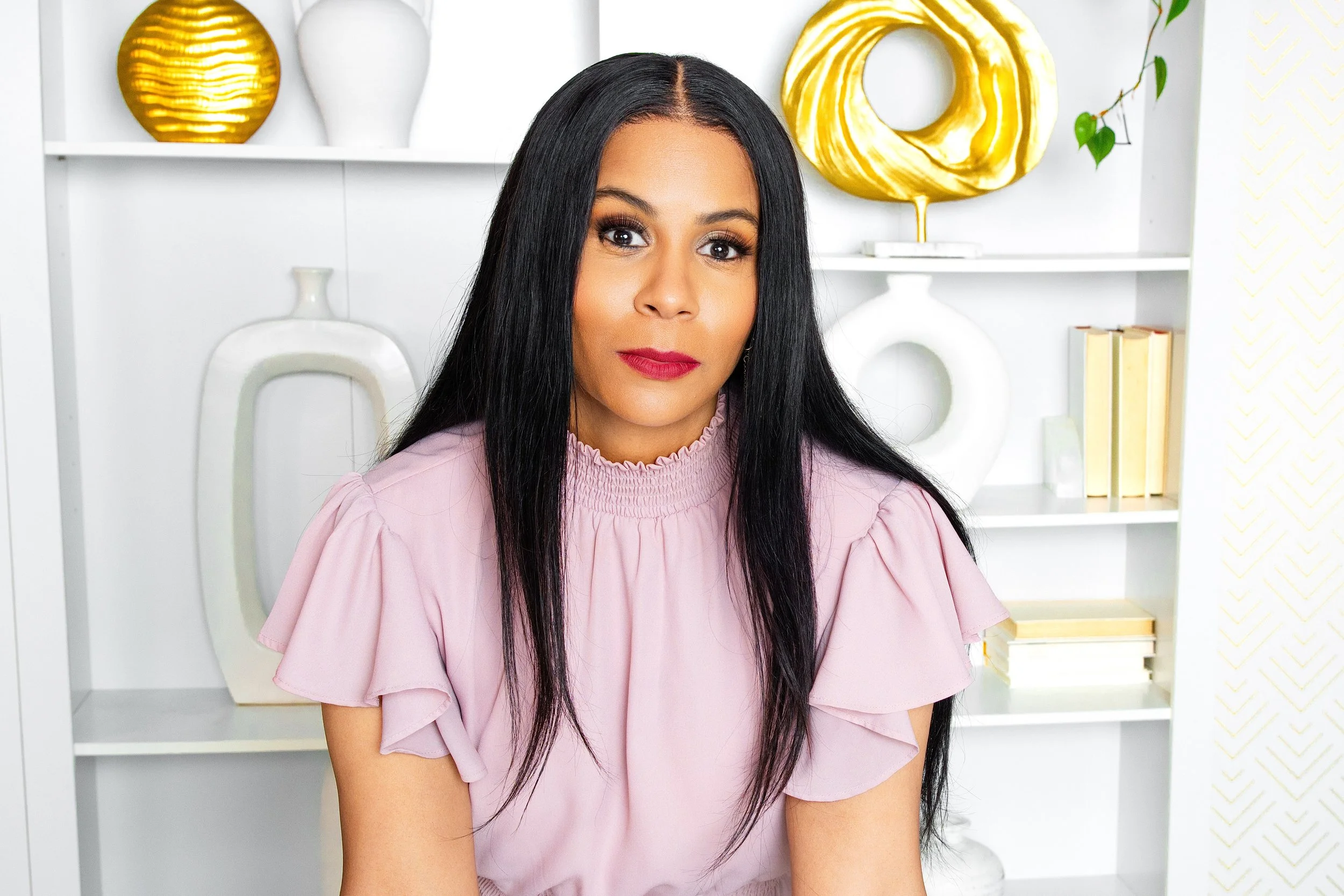 A woman with long black hair, wearing a pink ruffled blouse, sitting in front of a white bookshelf with decorative items, including gold and white vases and a stack of books.