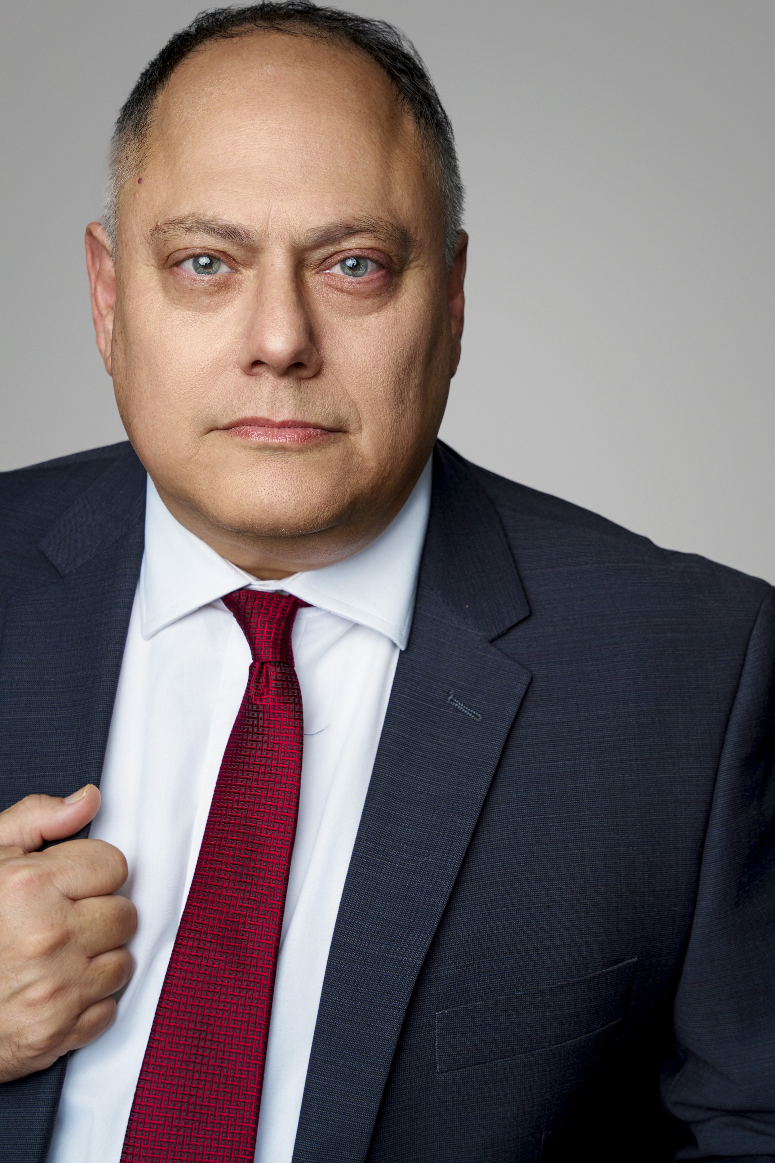 A middle-aged man wearing a dark suit, white dress shirt, and red tie, adjusting his jacket with a serious expression, against a plain gray background.