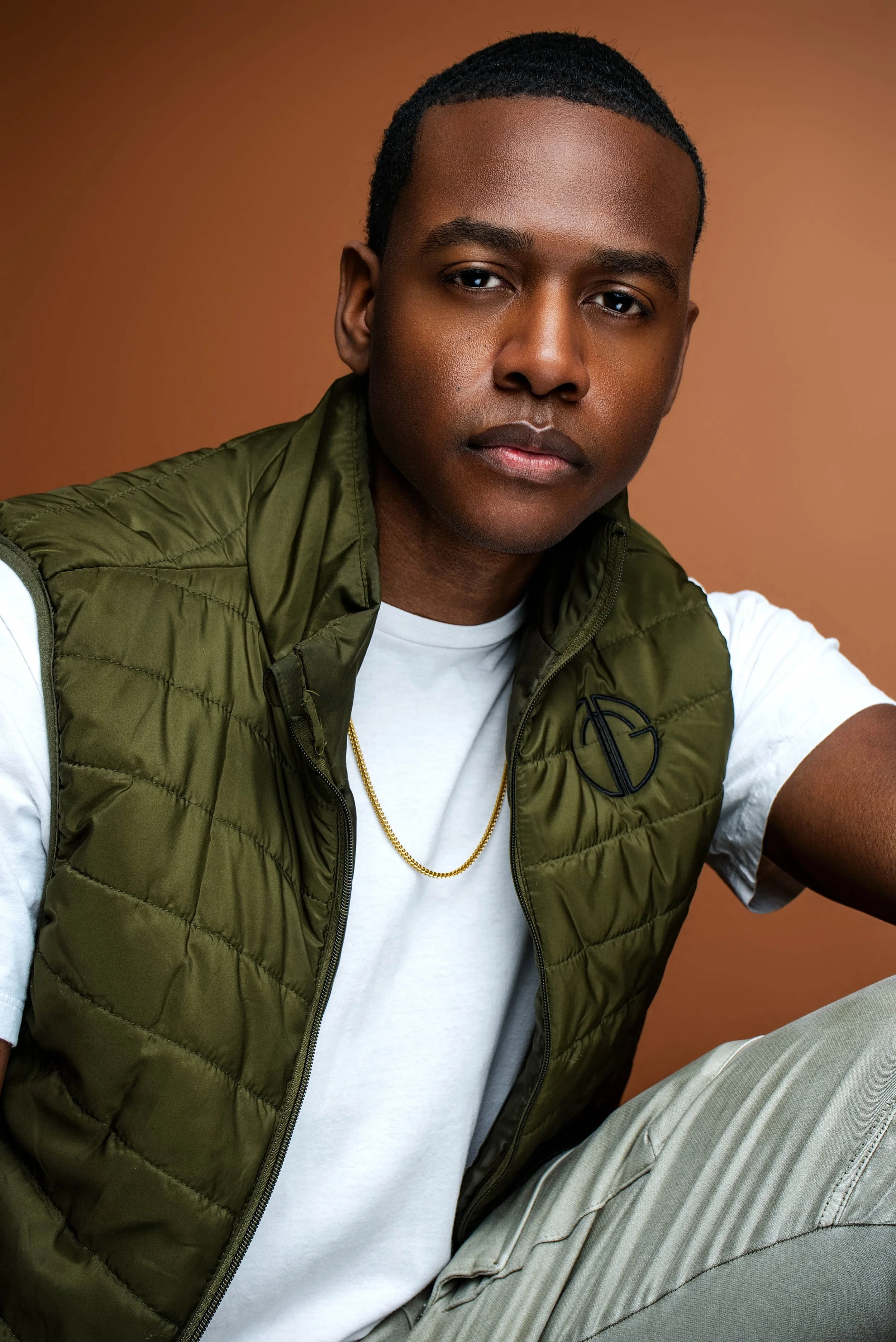 A young man with short black hair and medium skin tone wearing a white t-shirt, an olive green vest, a gold chain, and beige pants, sitting against a plain brown background.