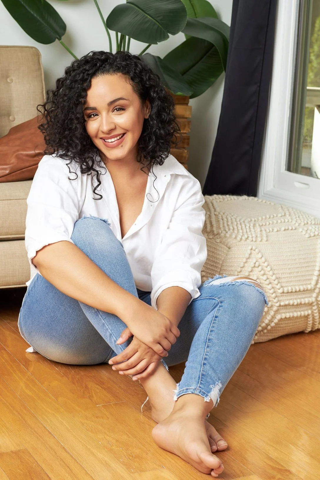 A young woman with curly hair smiling, sitting on a wooden floor with her legs crossed. She is wearing a white shirt and ripped jeans, near a large green plant and a window.