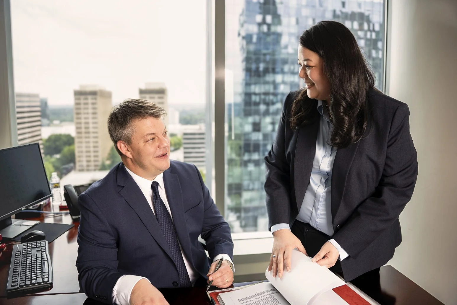 A man and woman in professional attire having a discussion in an office with a city view outside the window.