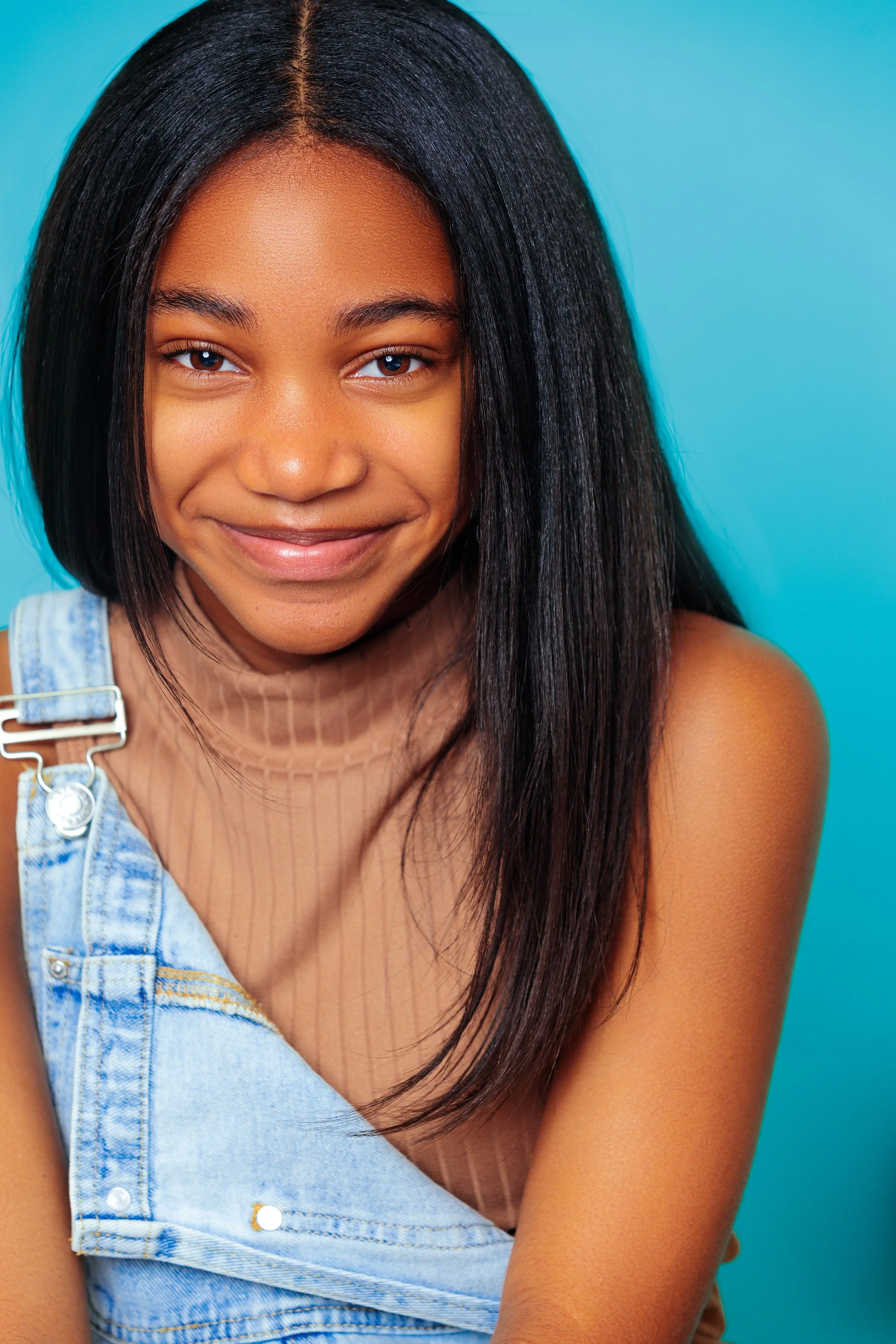 A young woman with long black hair, wearing a beige turtleneck top and denim overalls, smiling against a blue background.