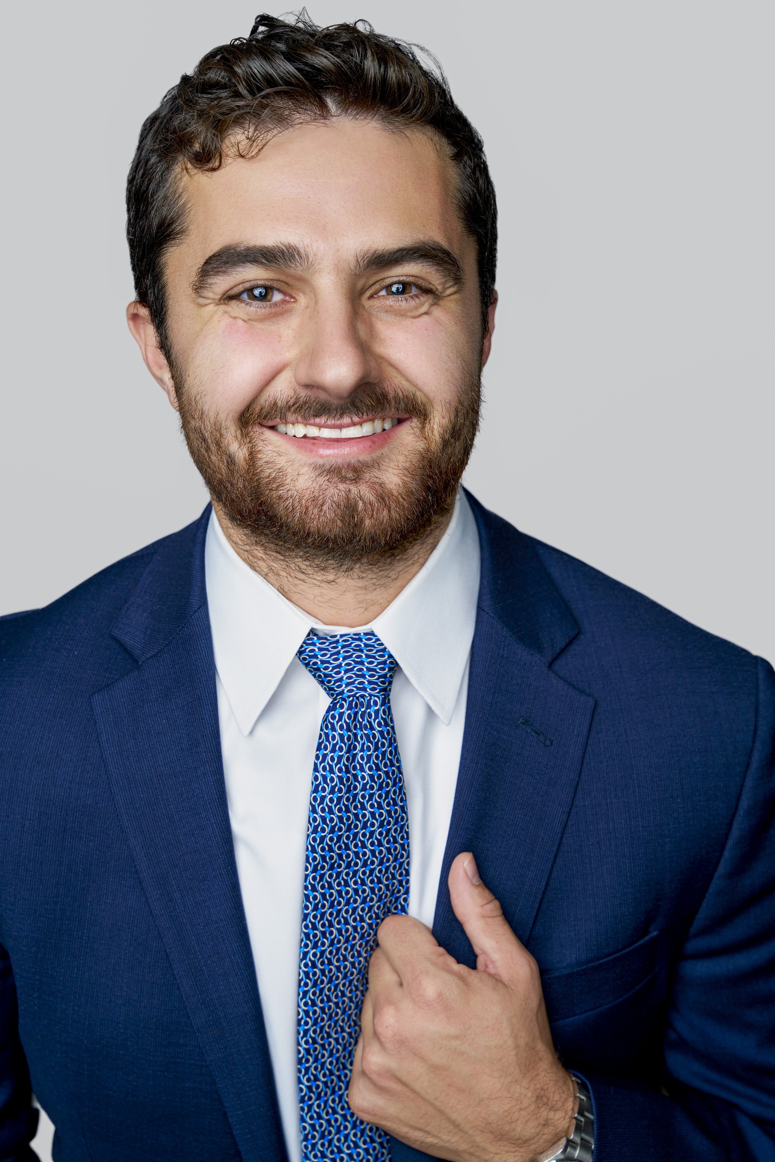 Headshot of a man smiling, wearing a navy suit, white shirt, and blue patterned tie, with a gray background.