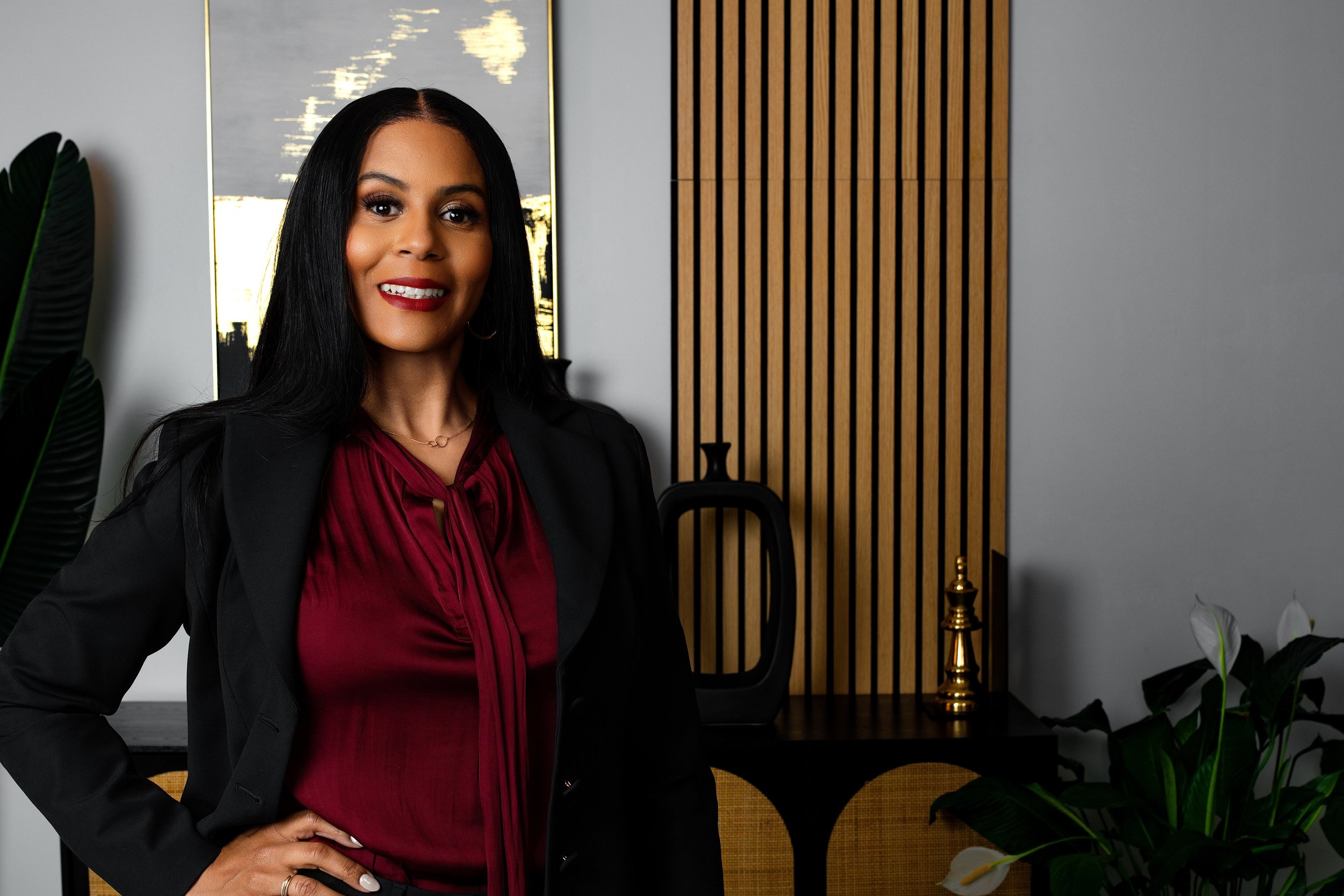 A woman with long black hair wearing a burgundy blouse and black blazer standing in an office environment with plants and decorative objects in the background.