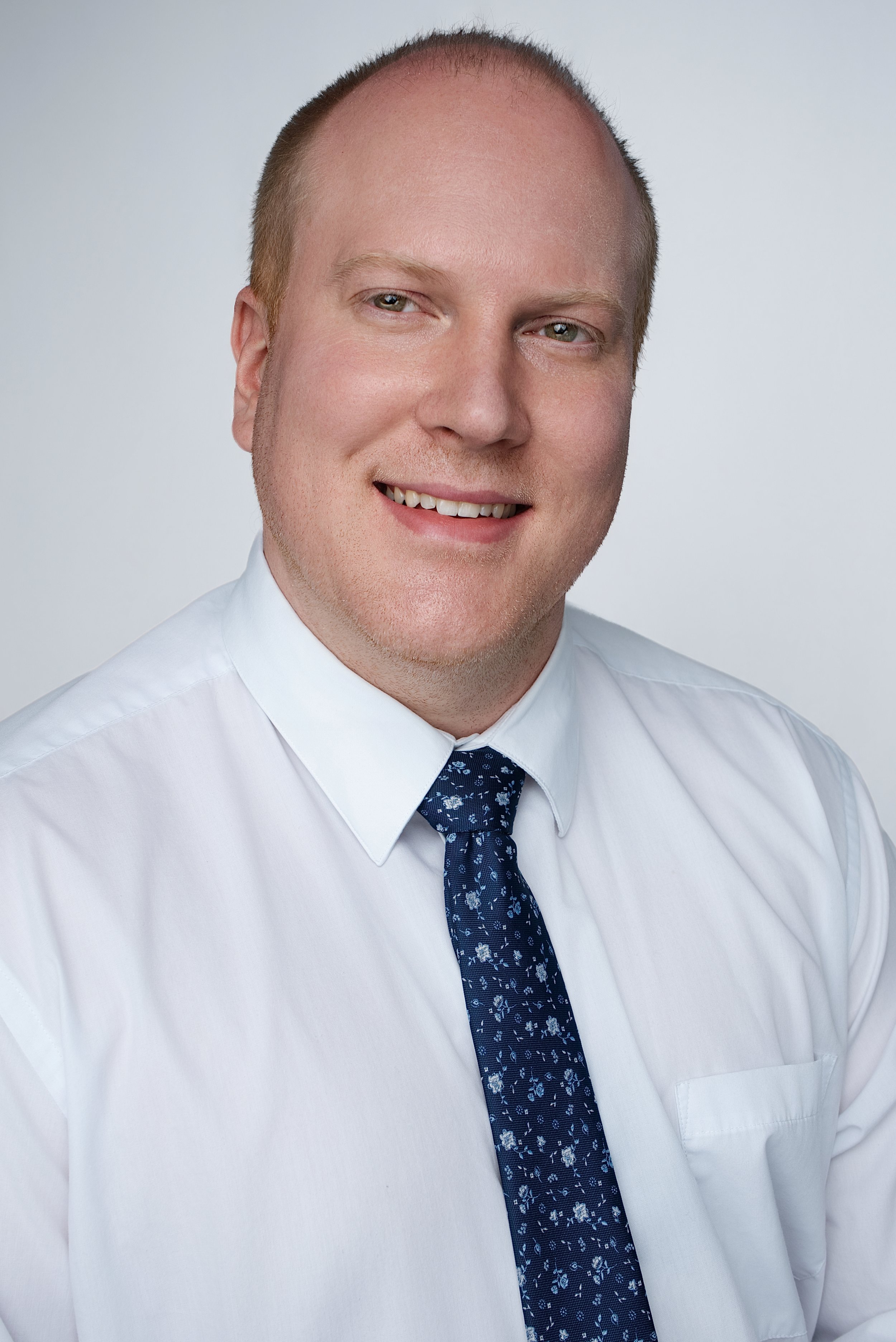 Professional headshot of a smiling man in a white shirt and dark blue floral tie against a light background.
