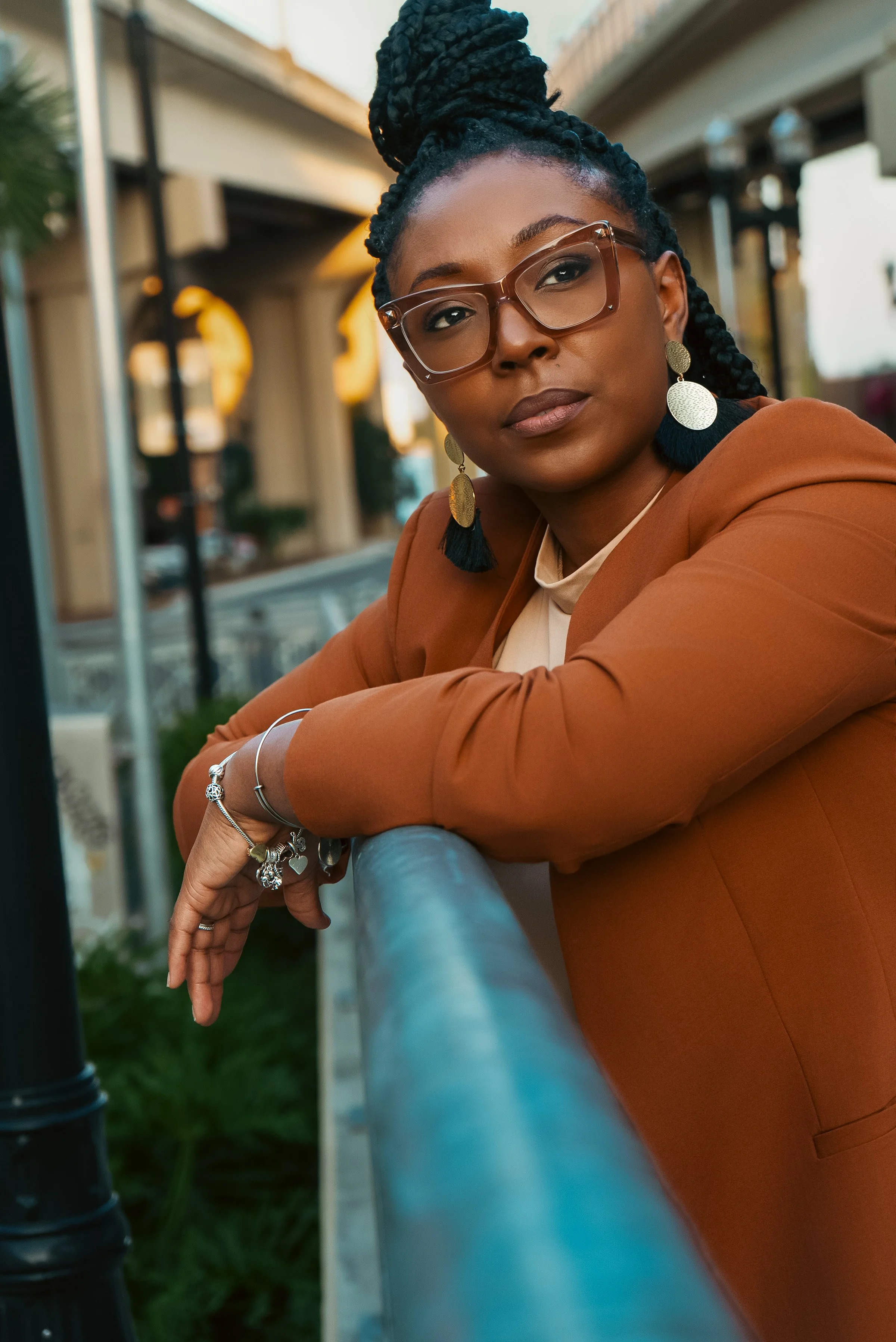 A woman with braided hair and glasses leaning on a railing outdoors.