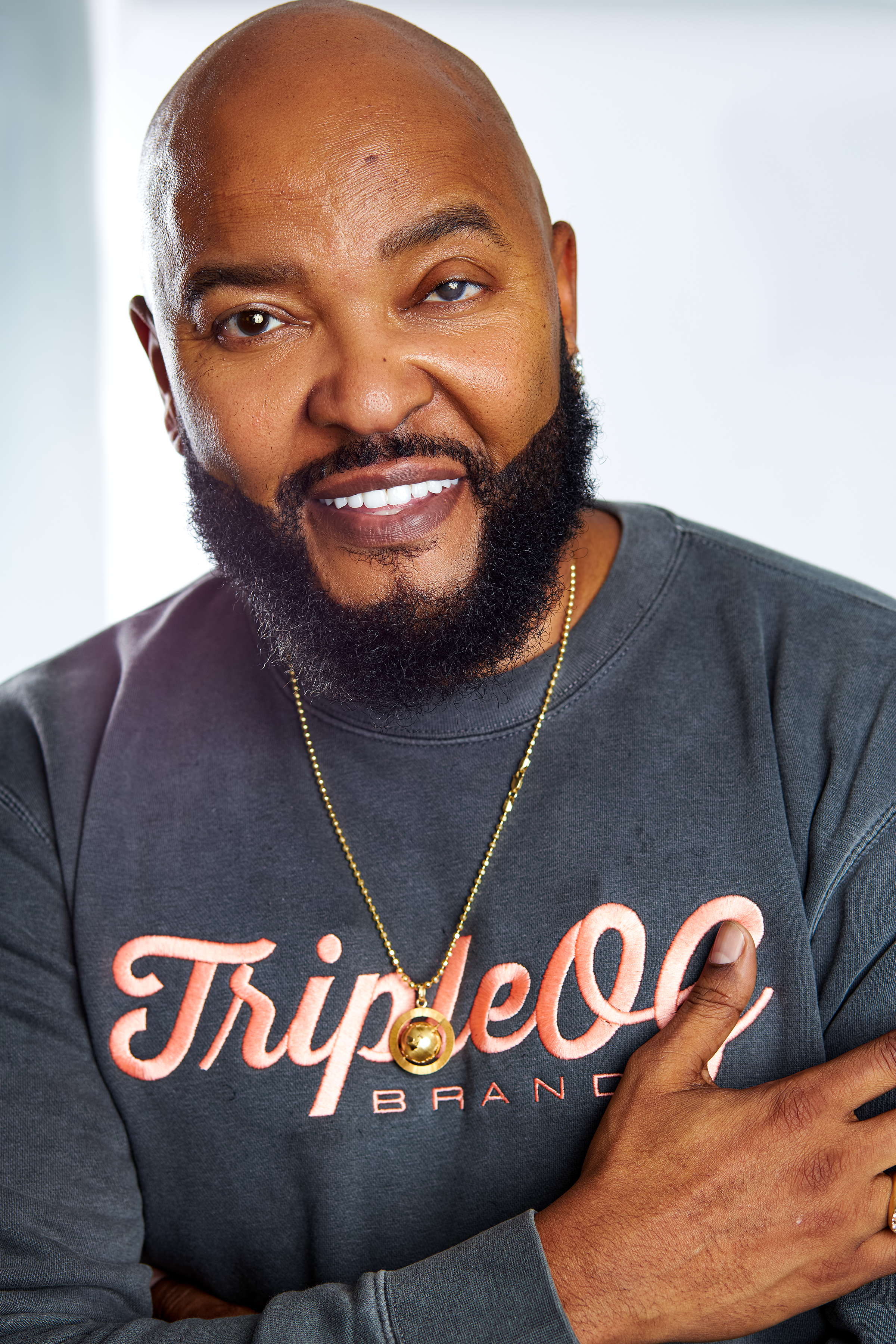 A smiling man with a bald head and beard, wearing a gray sweatshirt with the words 'Triple OG' and 'Brand' on it, and a gold necklace.