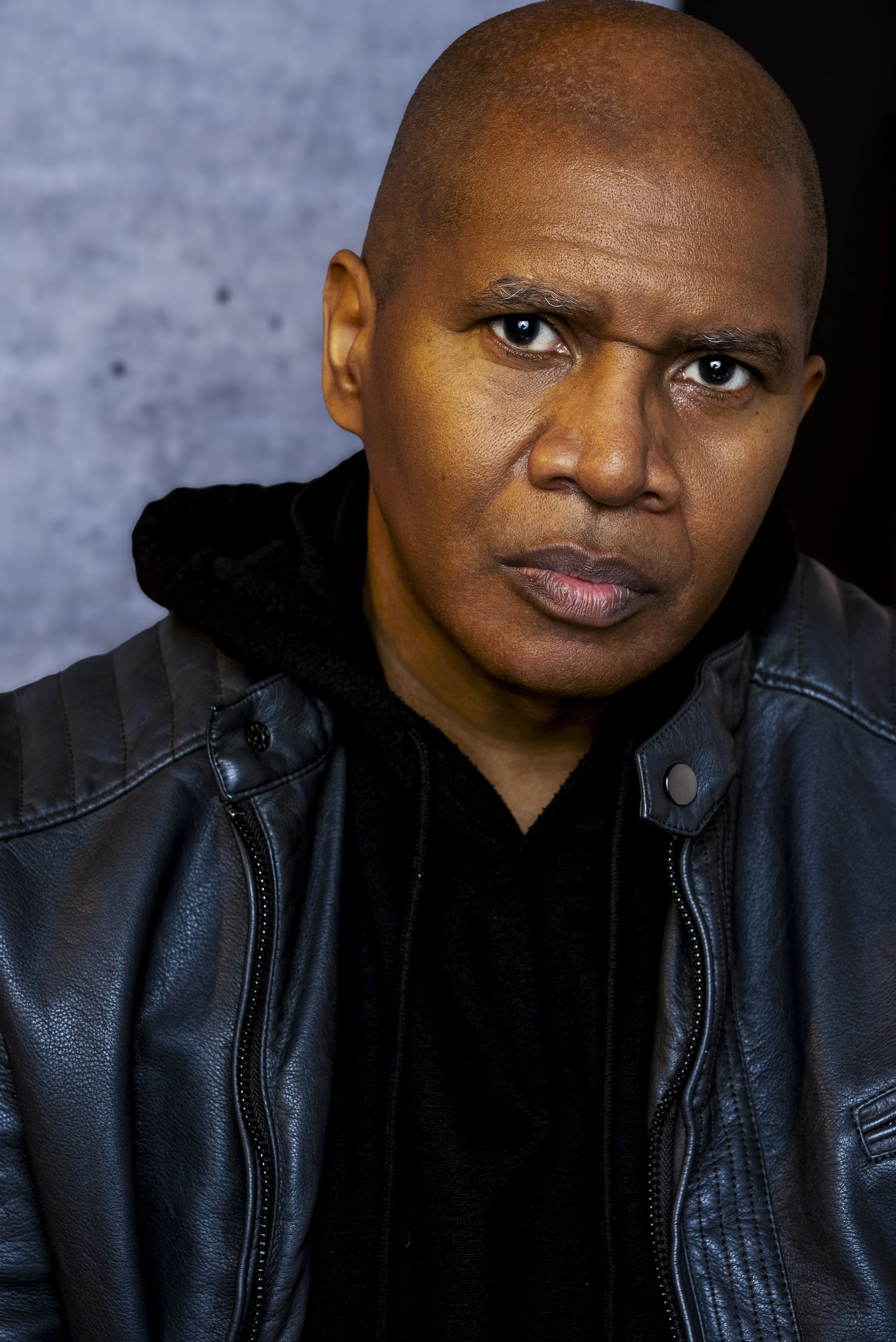Close-up of a serious man with a shaved head wearing a black leather jacket and black hoodie, looking directly at the camera against a textured background.