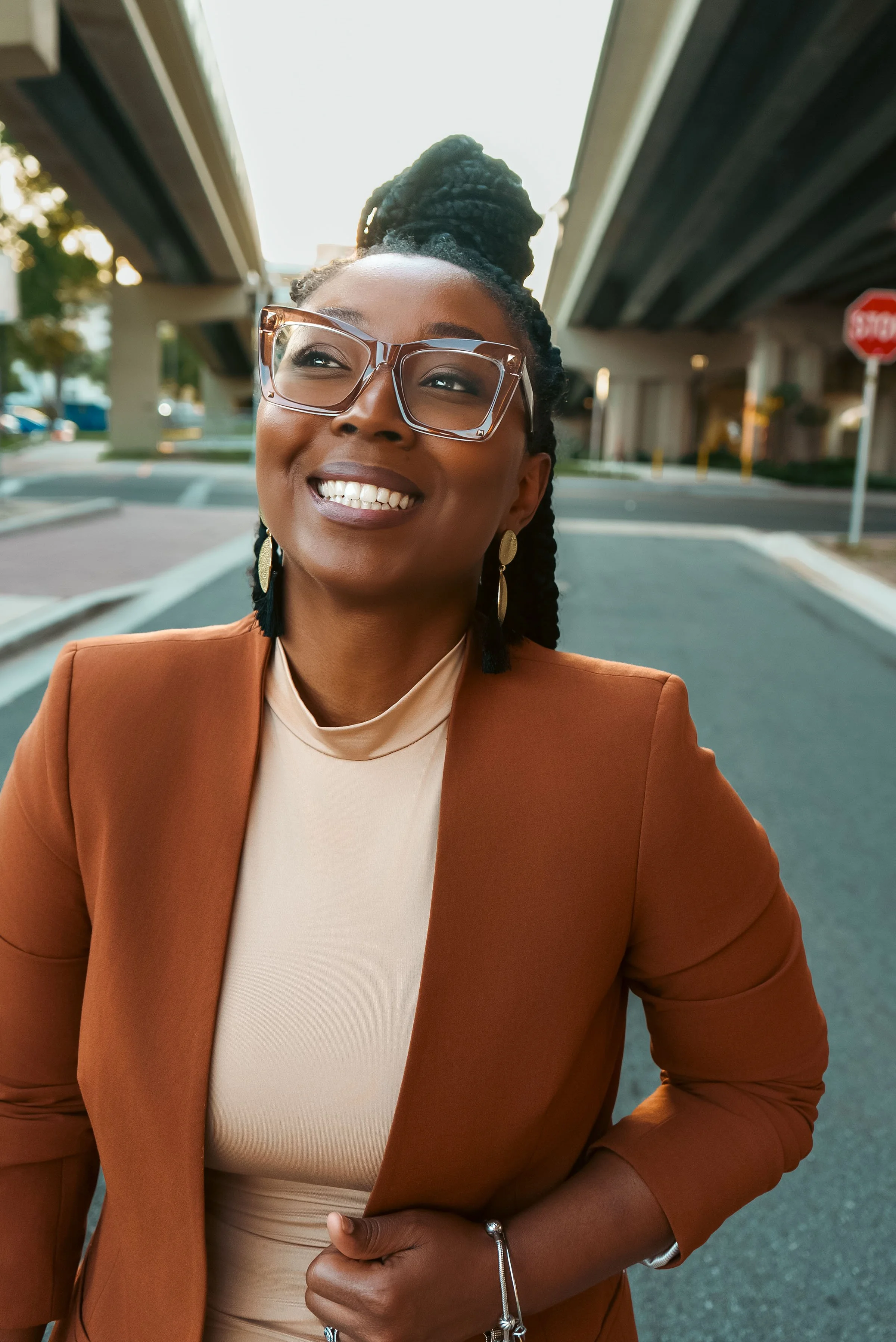 Woman smiling outdoors under a highway overpass, wearing glasses, earrings, a beige top, and a brown blazer.