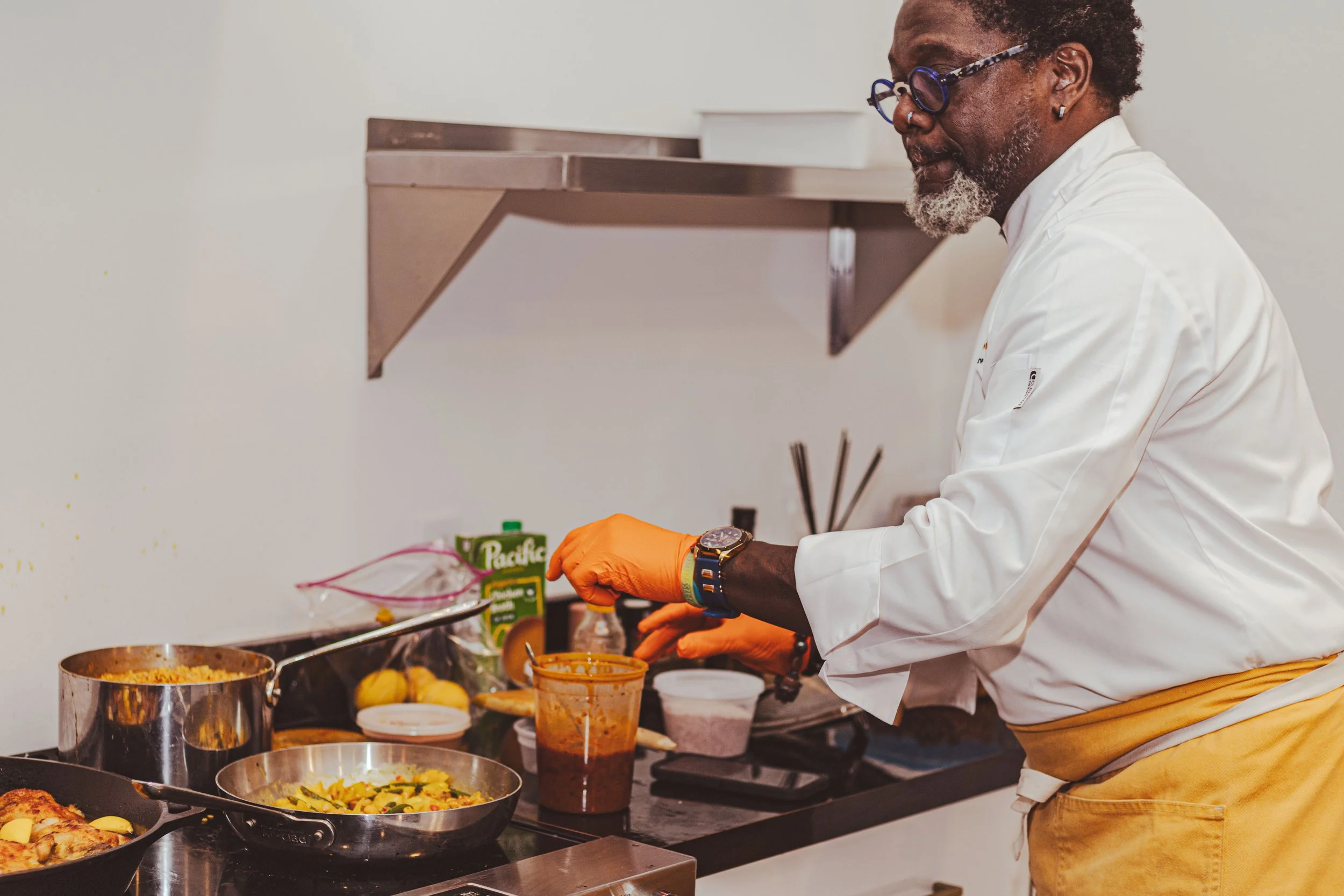 A man in a white chef's coat and orange gloves preparing food in a kitchen with various ingredients and cooking utensils on the countertop.