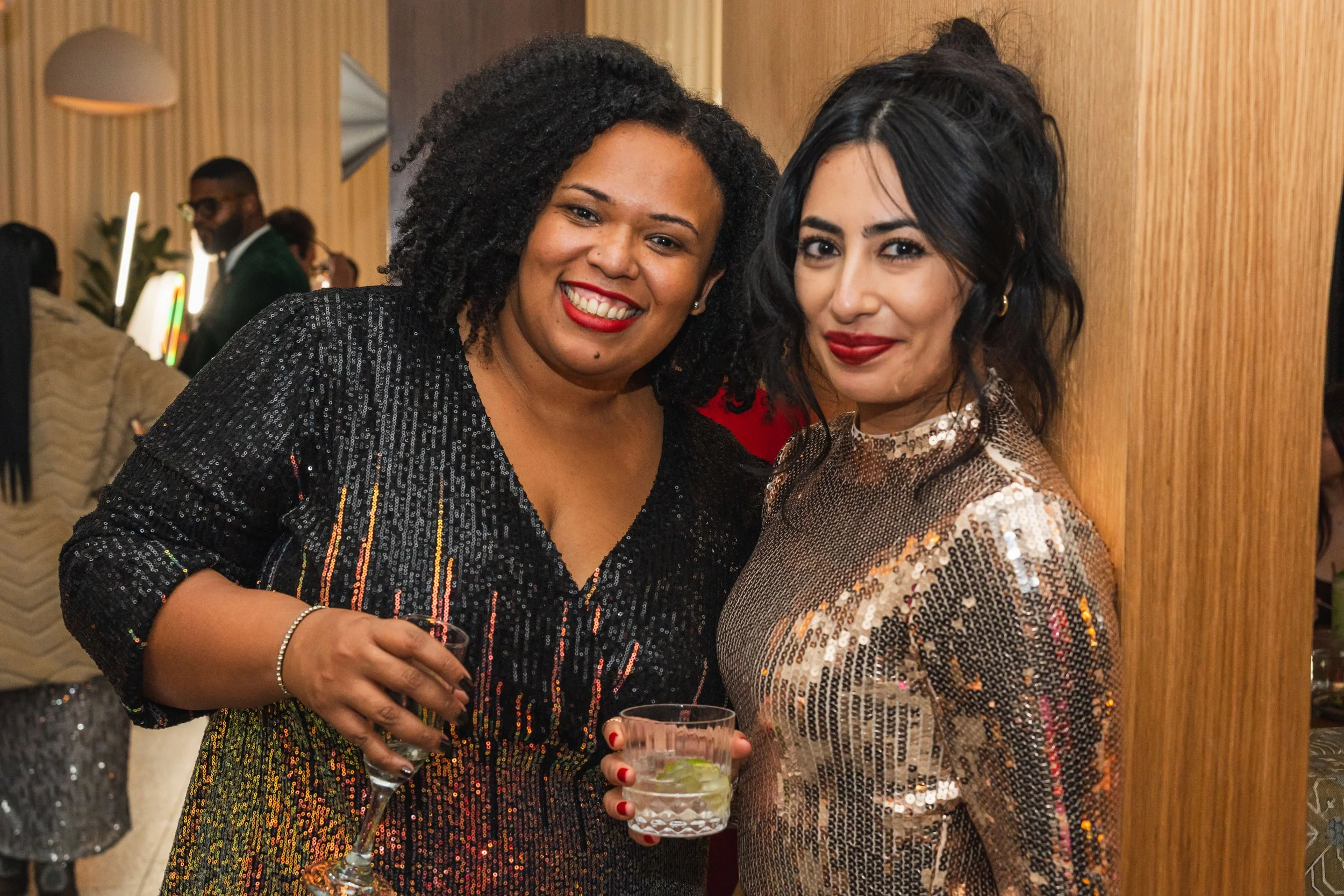 Two women dressed in shiny, sequined outfits smiling and holding drinks at a party or social gathering.