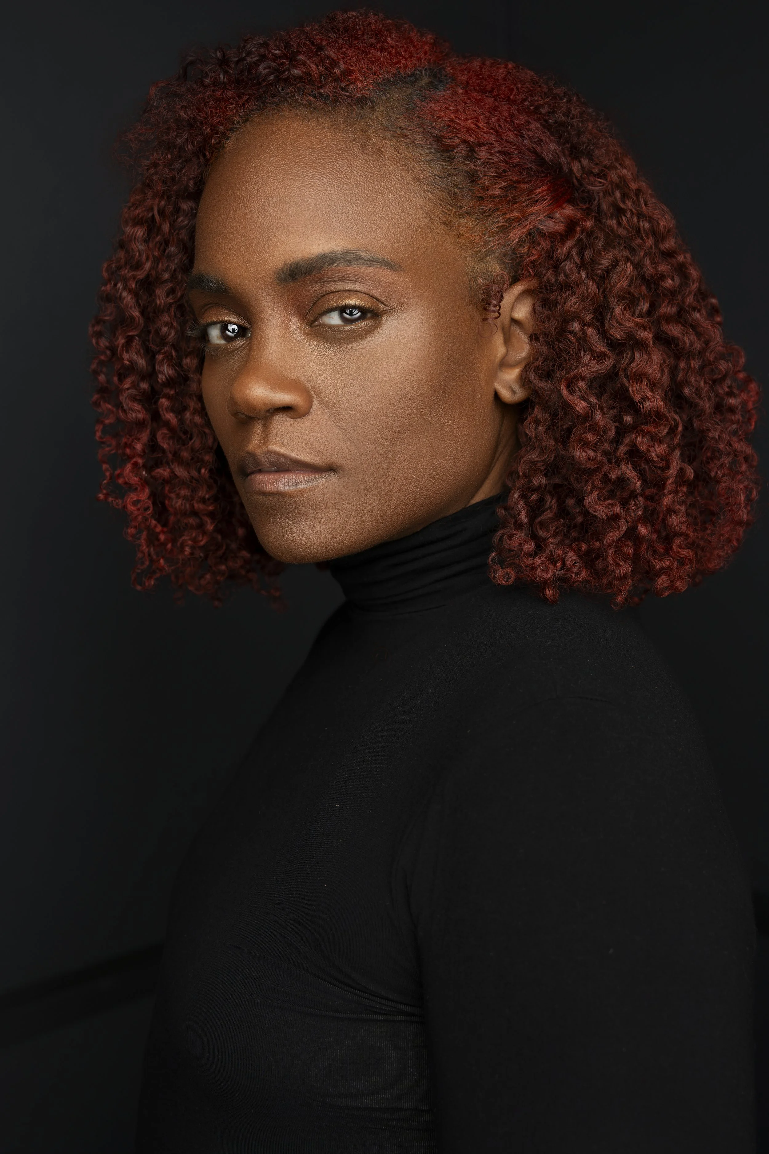Portrait of a woman with curly red hair wearing a black turtleneck against a dark background.