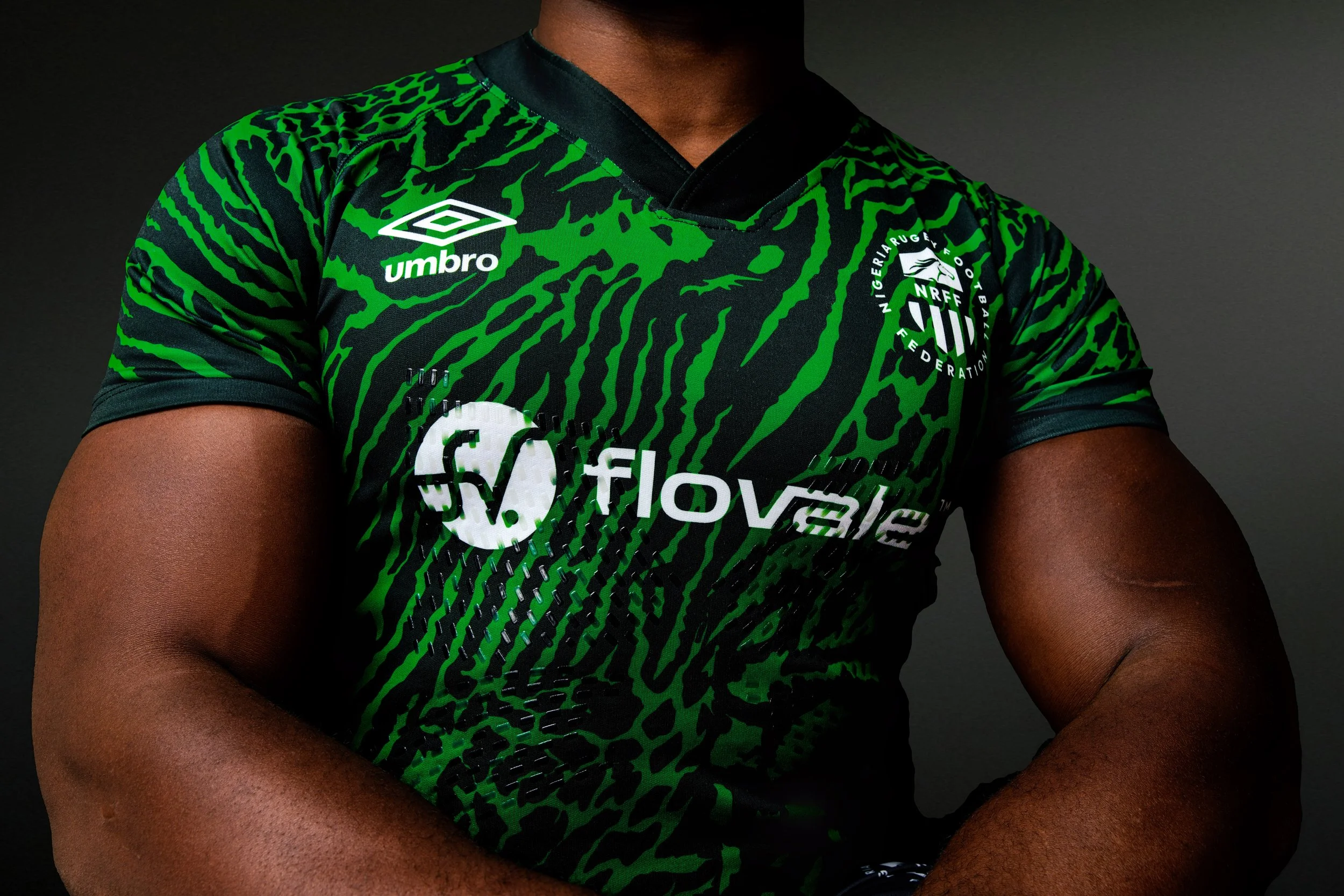 Close-up of a muscular person wearing a green and black sports jersey with various logos, including Umbro, SoFive, and a Nigerian rugby federation emblem, against a dark background.