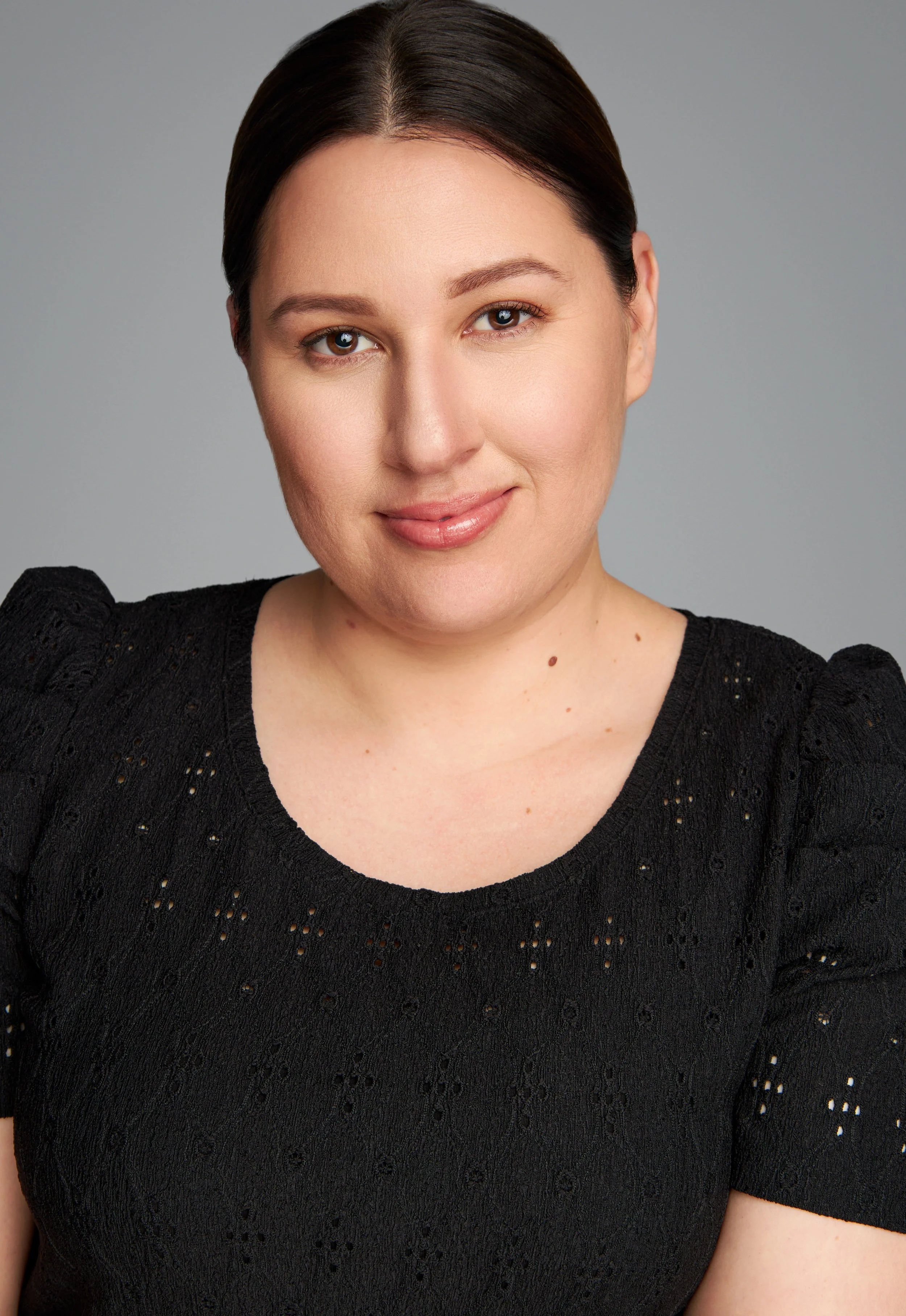 A woman with dark brown hair styled in a middle part, wearing a black textured top, smiling softly against a neutral gray background.