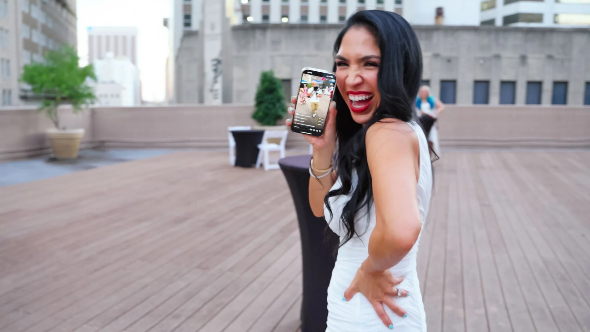 A woman with long dark hair and bright red lipstick is smiling and holding a smartphone, taking a selfie on a rooftop patio with wooden flooring and modern buildings in the background.