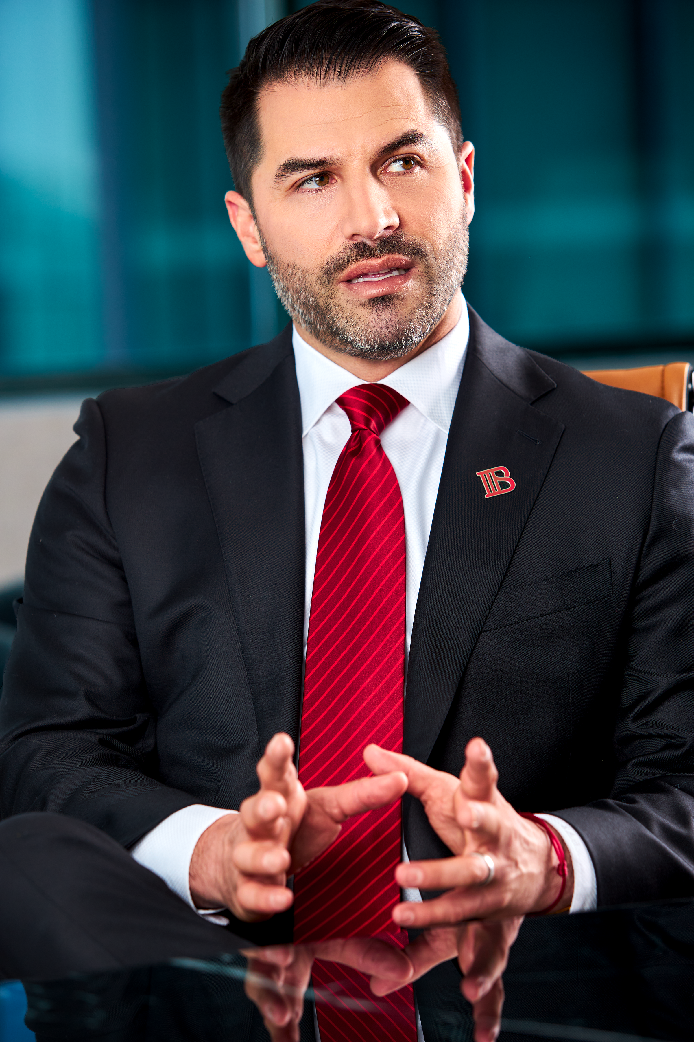 A man in a black suit with a white shirt and a red striped tie, sitting at a table in a modern office, gesturing with his hands, and wearing a lapel pin with a red and black logo.