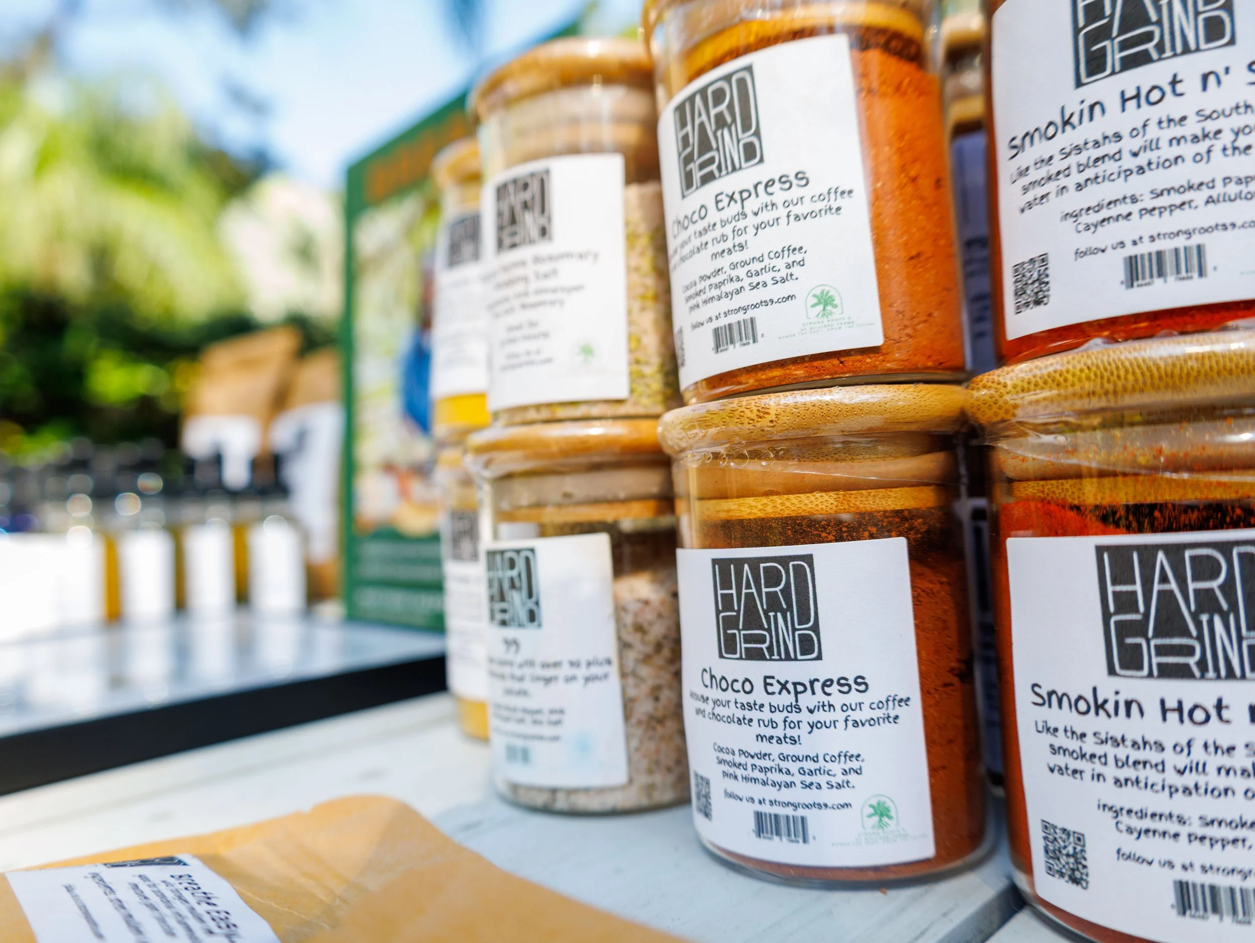 Multiple jars of spice blends labeled "Hard Grind," including "Choco Express" and "Smokin Hot n' Sweet" on an outdoor market stall, with a blurred background of trees and sky.