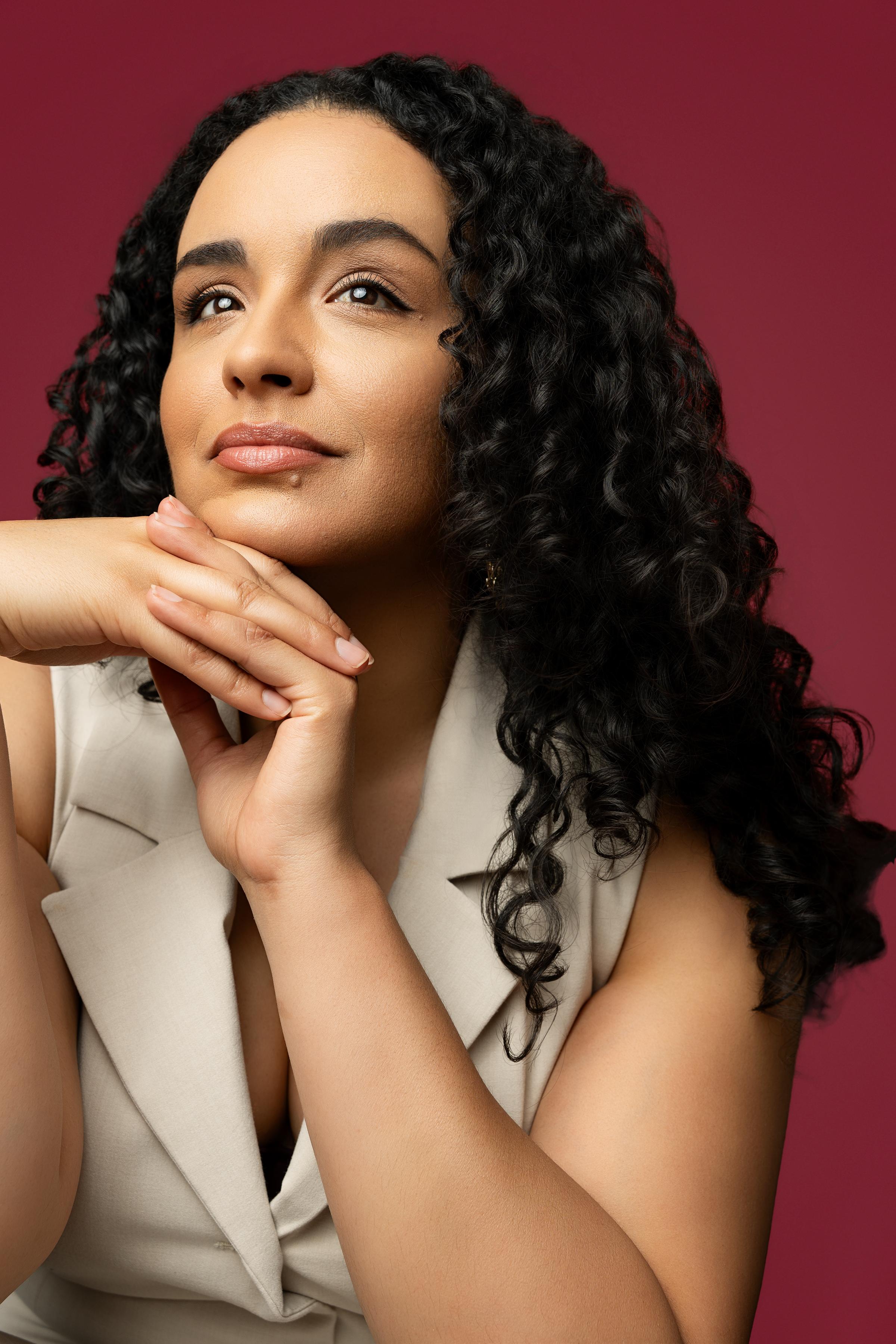 A woman with curly dark hair and light skin looking thoughtfully upward, resting her chin on her hands, against a maroon background.