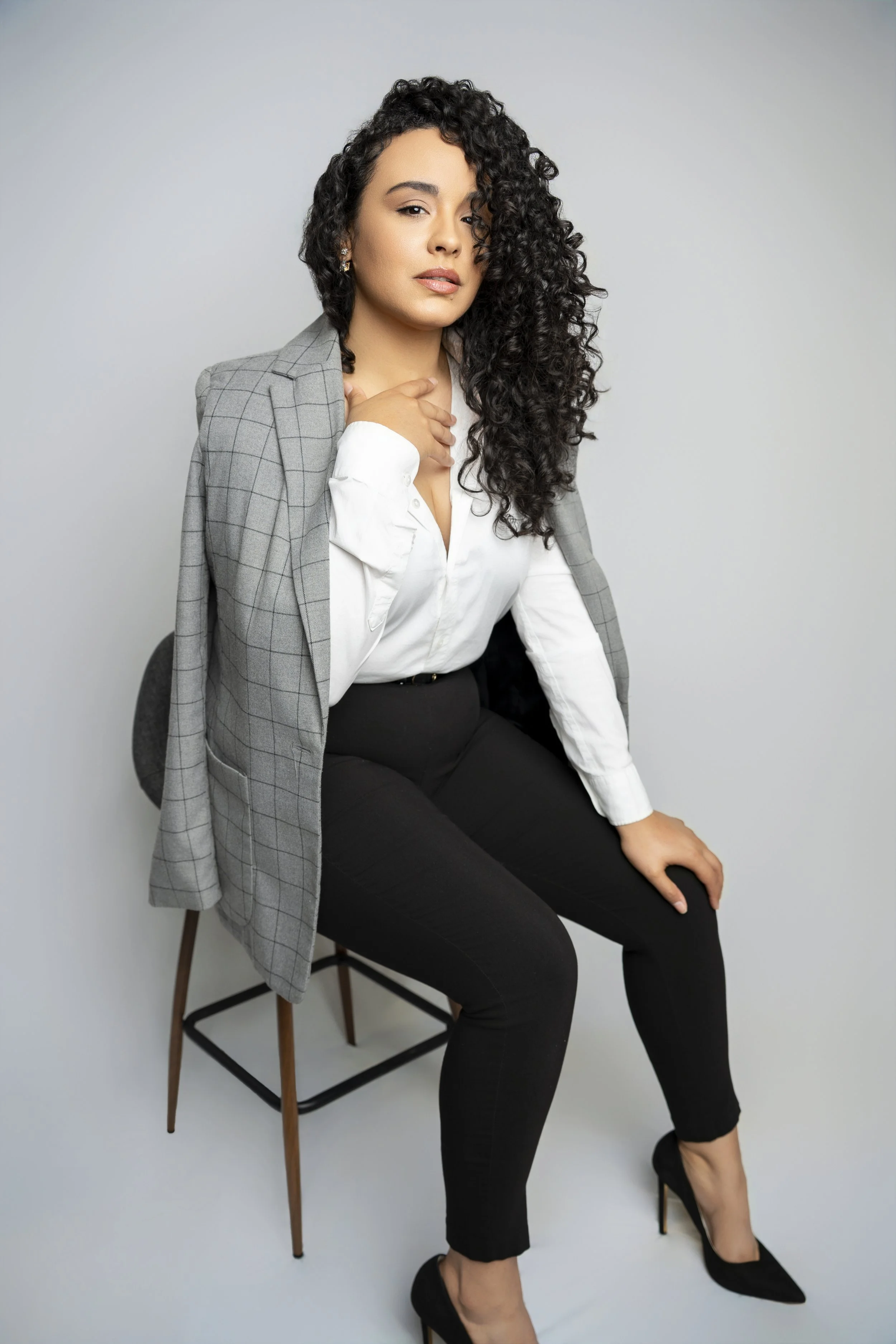 A woman with curly dark hair sitting on a chair against a plain gray background. She is wearing black pants, black heels, a white blouse, and a gray blazer draped over her shoulders, with her left hand on her thigh and her right hand touching her nec