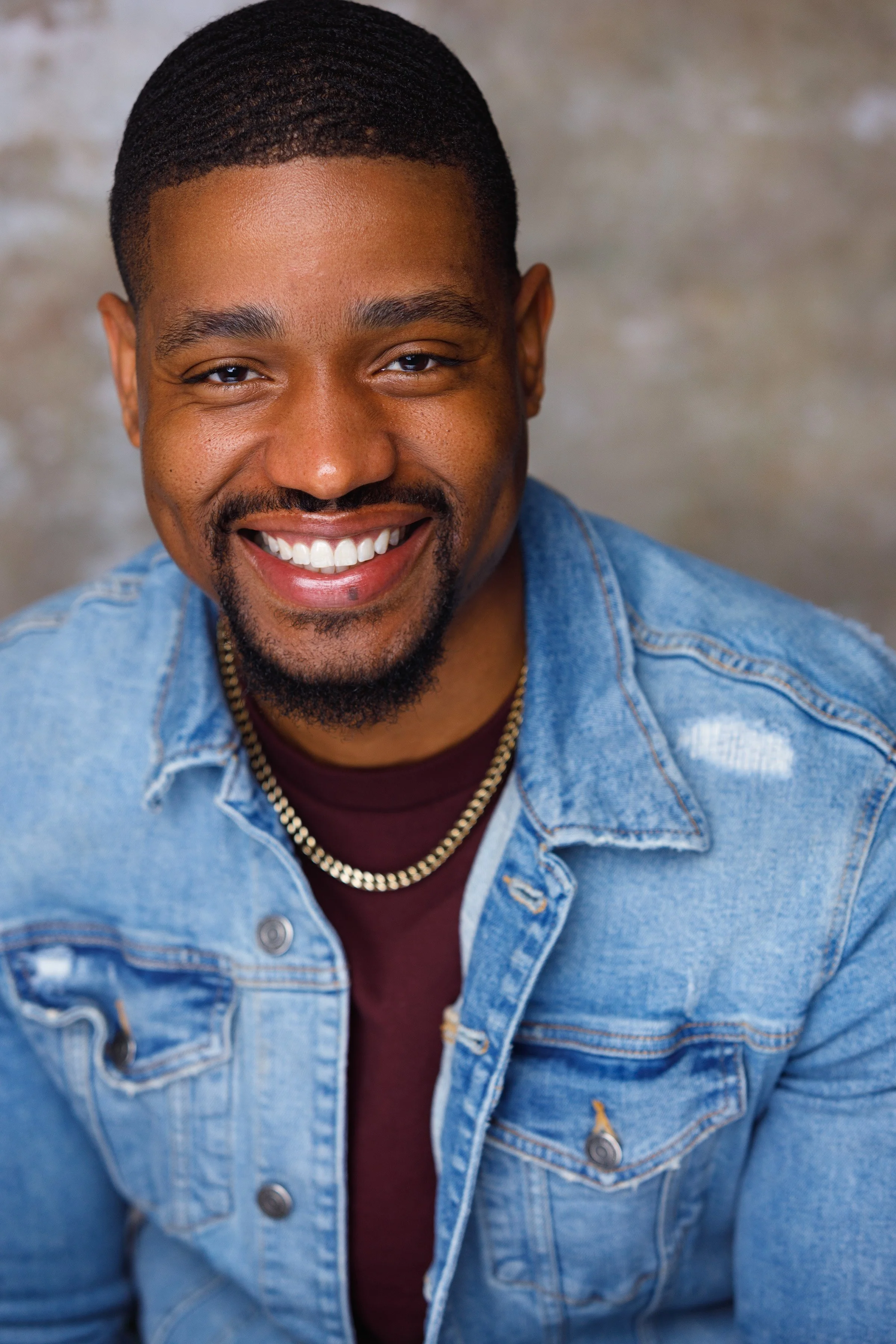 Portrait of a smiling man with dark skin, short hair, a goatee, wearing a denim jacket over a maroon shirt, a gold chain, and a warm expression.