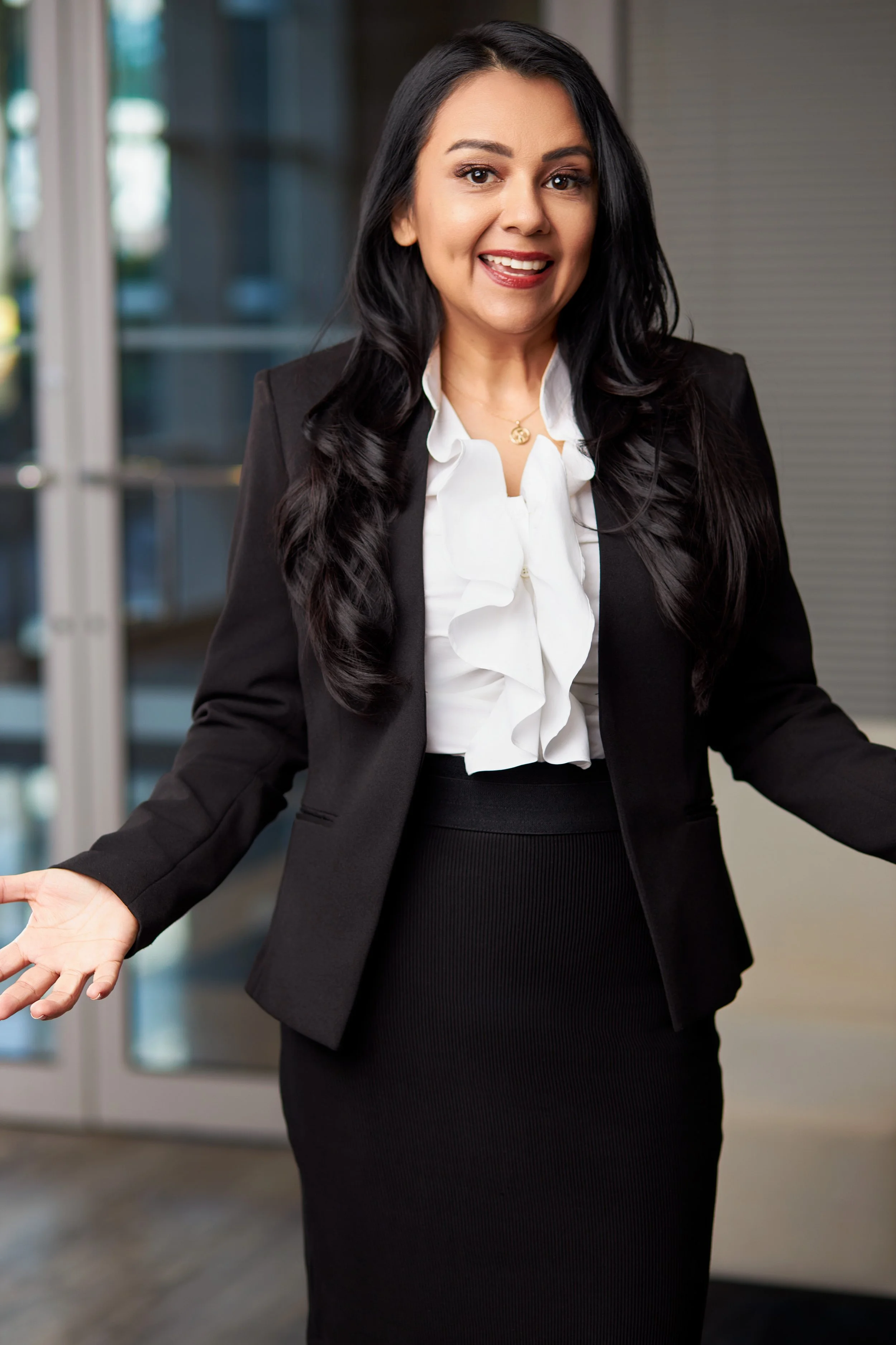 A woman with long dark hair wearing a black blazer and a white ruffled blouse, standing indoors with an open, welcoming gesture.