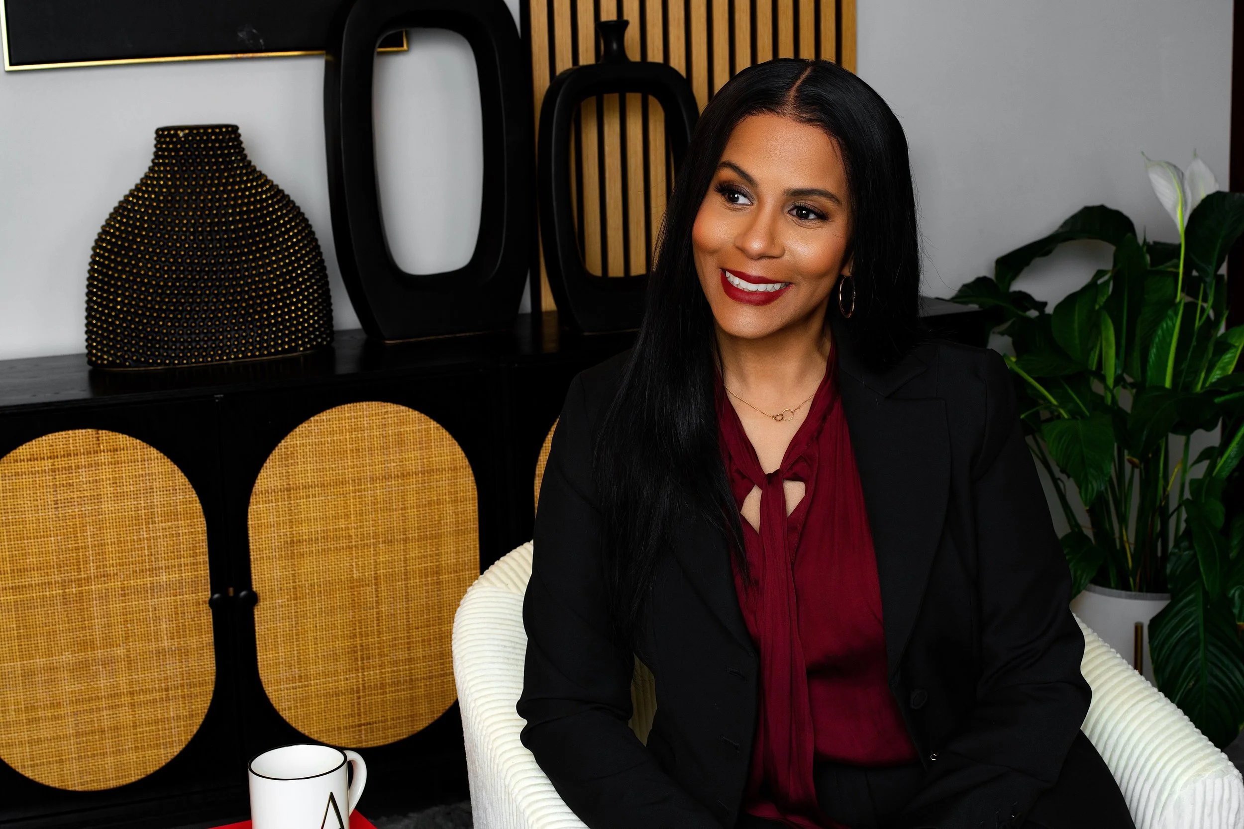 A woman with long black hair, wearing a black blazer and a deep red blouse, seated in a modern white armchair, smiling. Behind her is a black cabinet with decorative objects, including a black and gold textured vase and black geometric sculptures, an