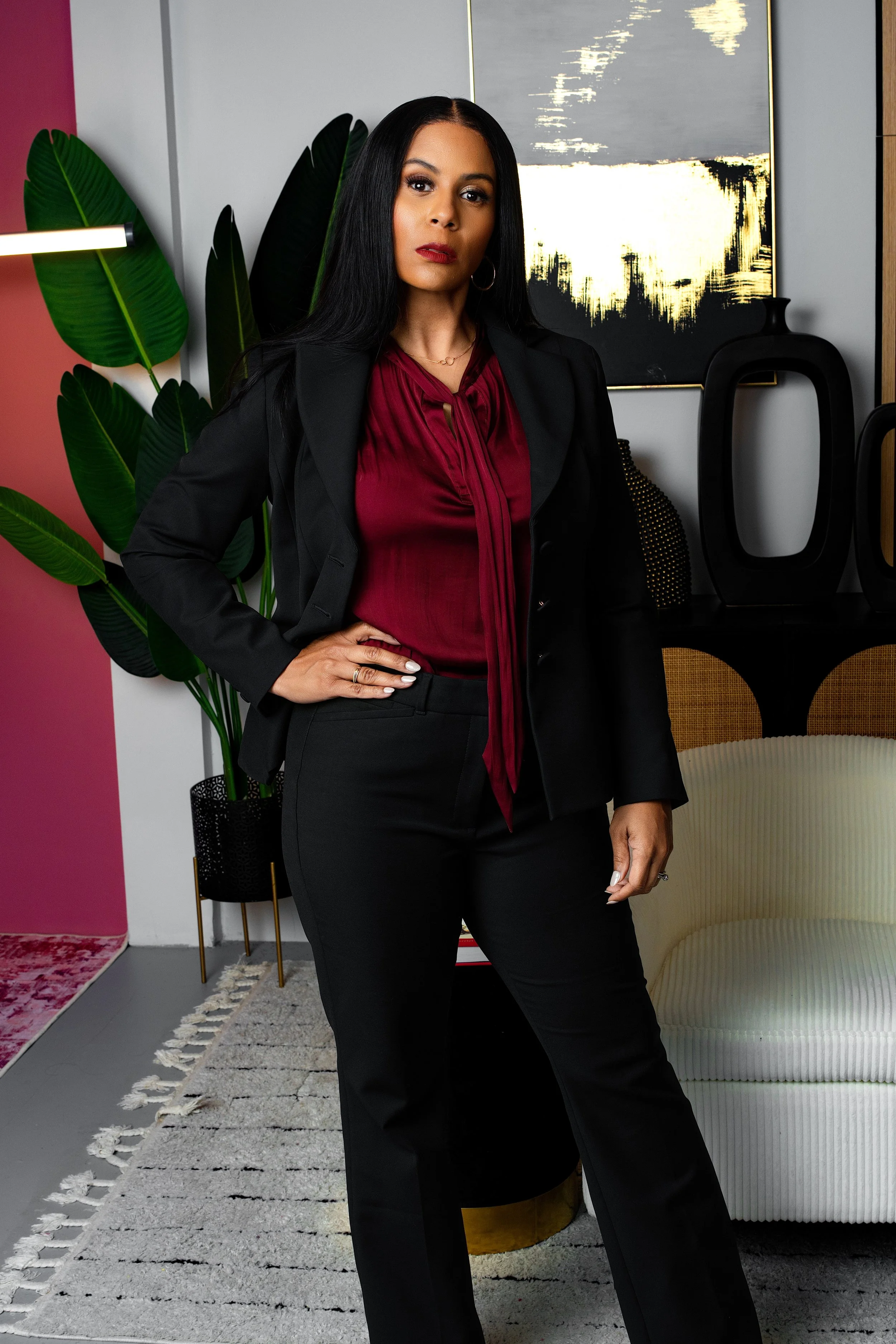 A woman with long black hair dressed in a black blazer and pants, and a red blouse, standing with her hand on her hip in a stylish modern living room with abstract artwork, large green plants, a white cushioned couch, decorative black vases, and a pa