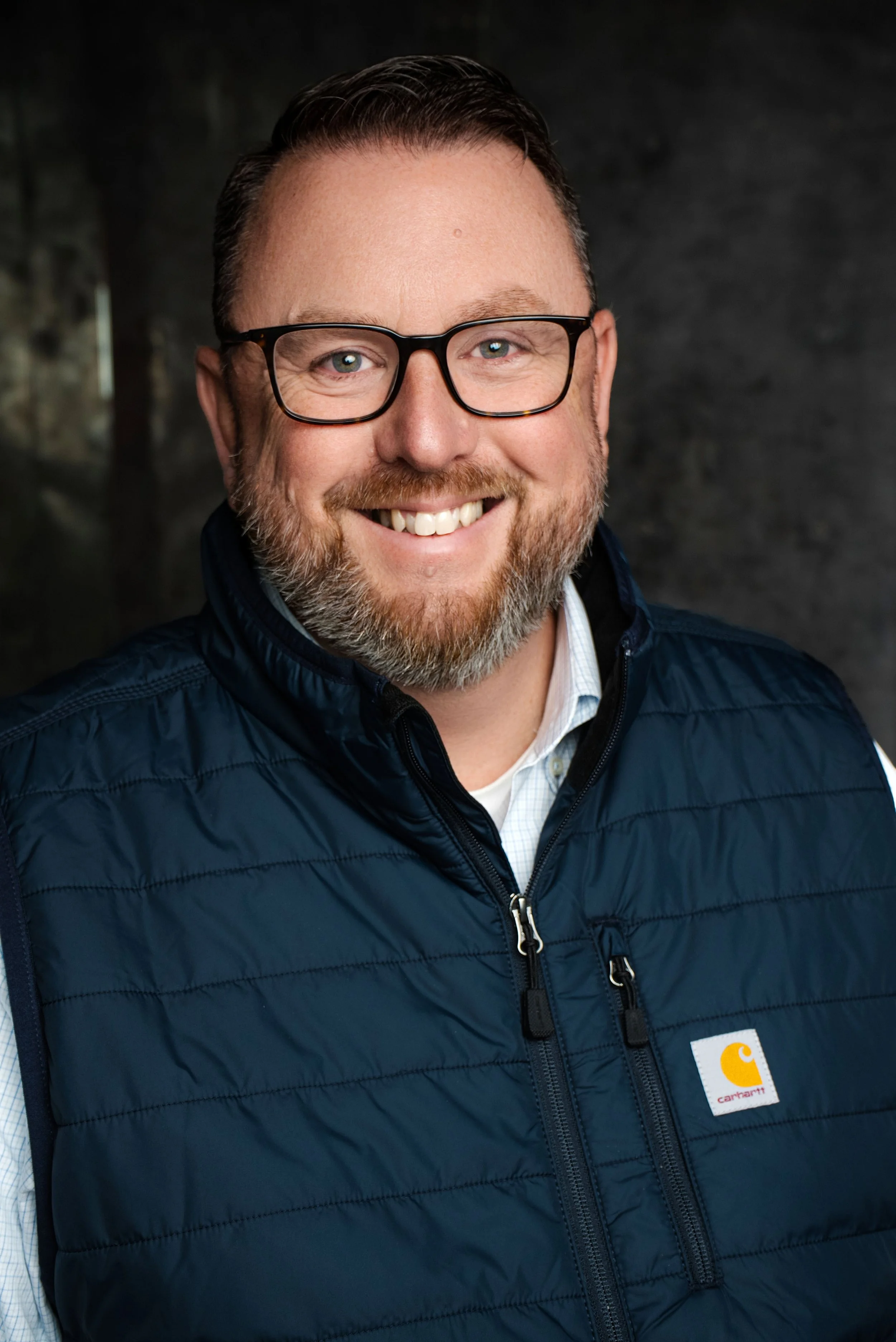 A smiling man with glasses and a beard wearing a navy blue Carhartt vest over a white shirt, posing indoors against a dark background.