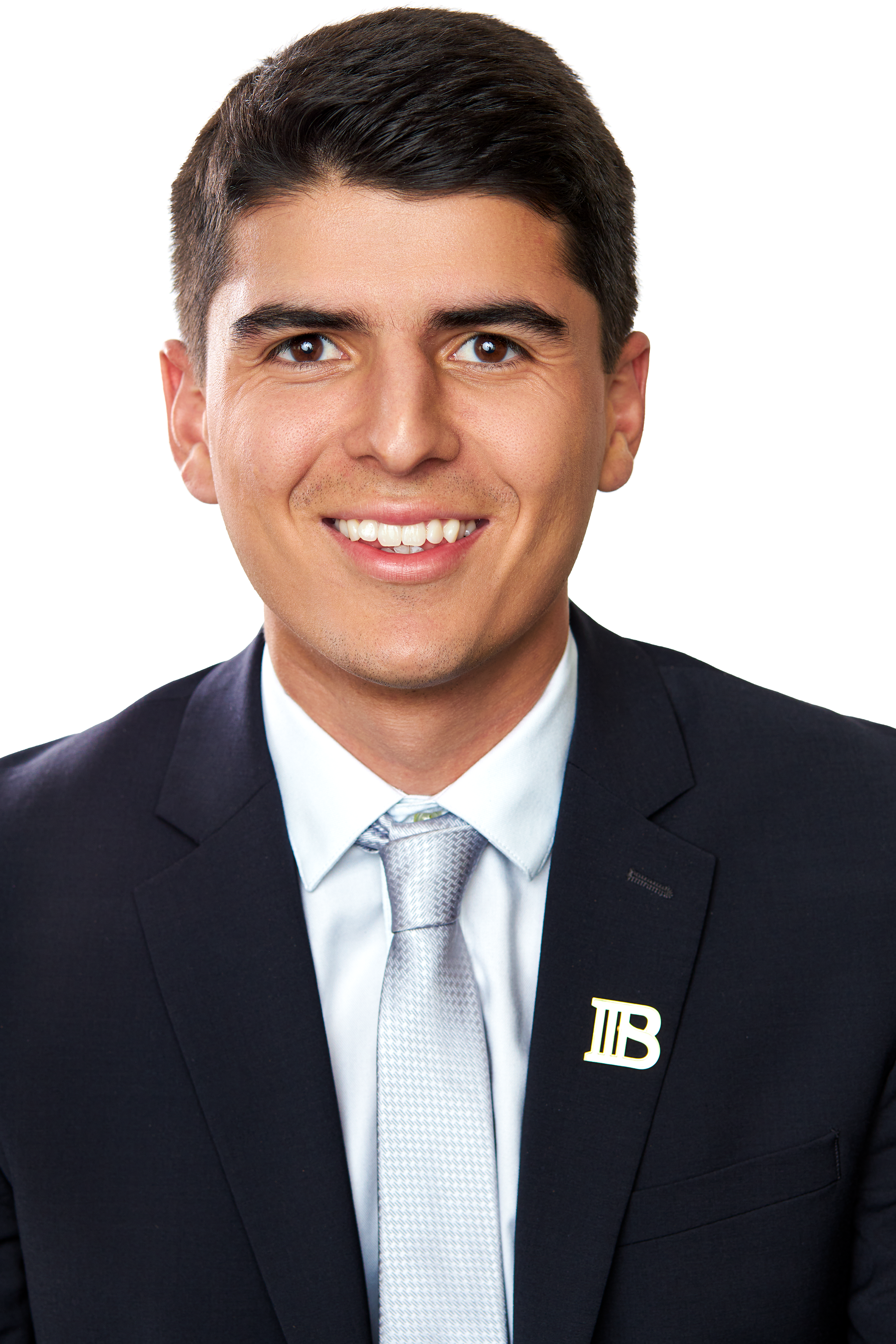 A young man in a suit and tie smiling, with a pin on his lapel that has two vertical lines and a horizontal line, against a plain white background.