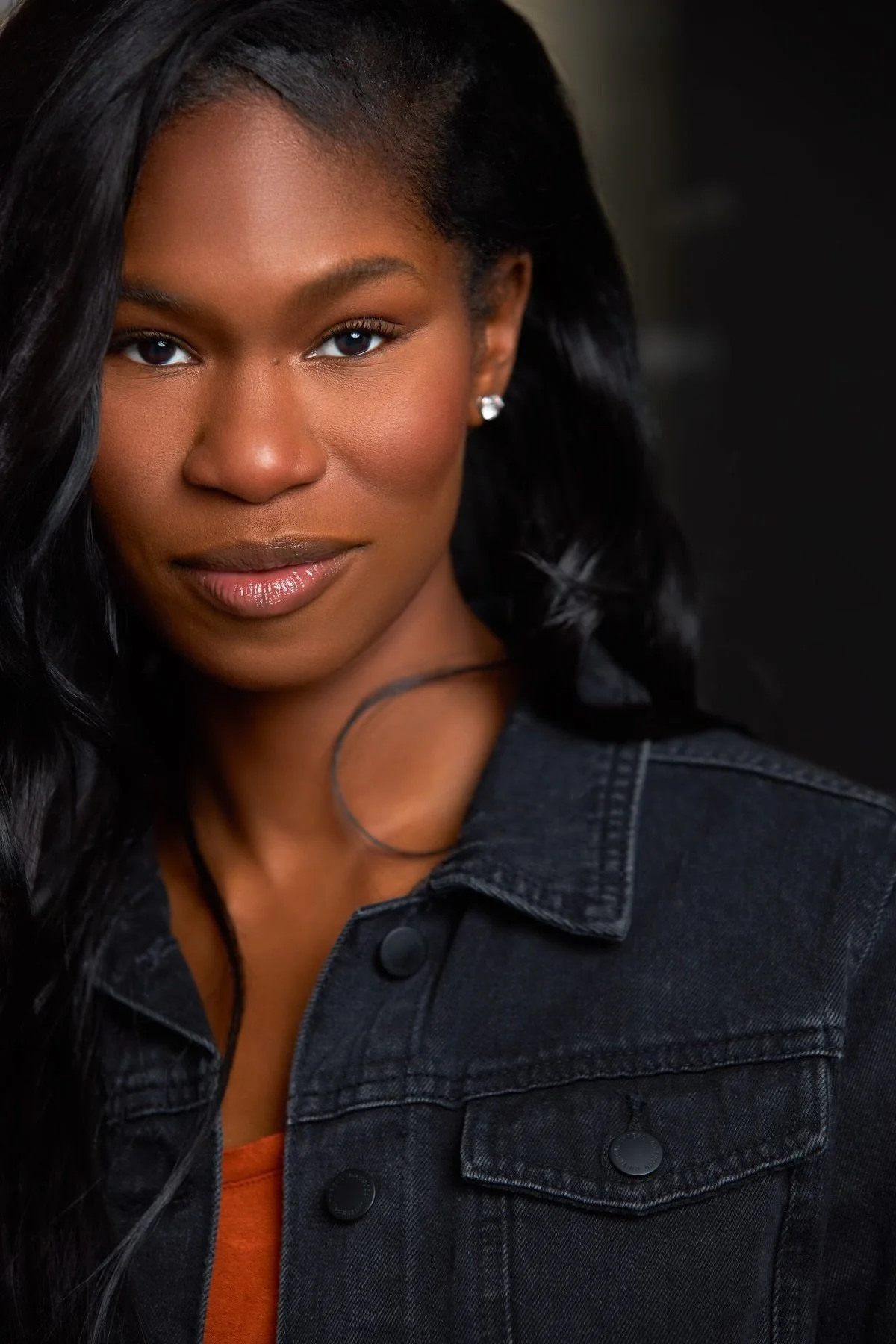 A woman with dark hair, wearing a black denim jacket and pearl earrings, posing with a confident expression against a dark background.