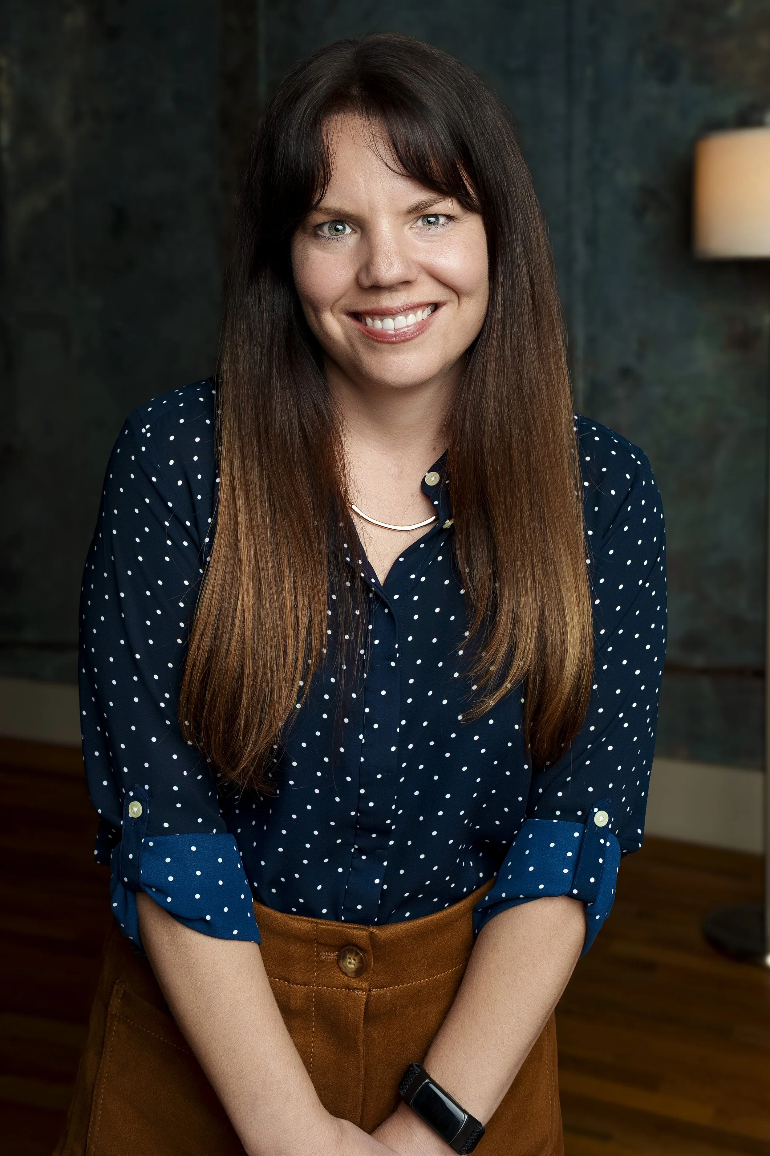 A woman with long brown hair wearing a navy blue polka dot blouse and a brown skirt, smiling indoors with a lamp and textured wall in the background.