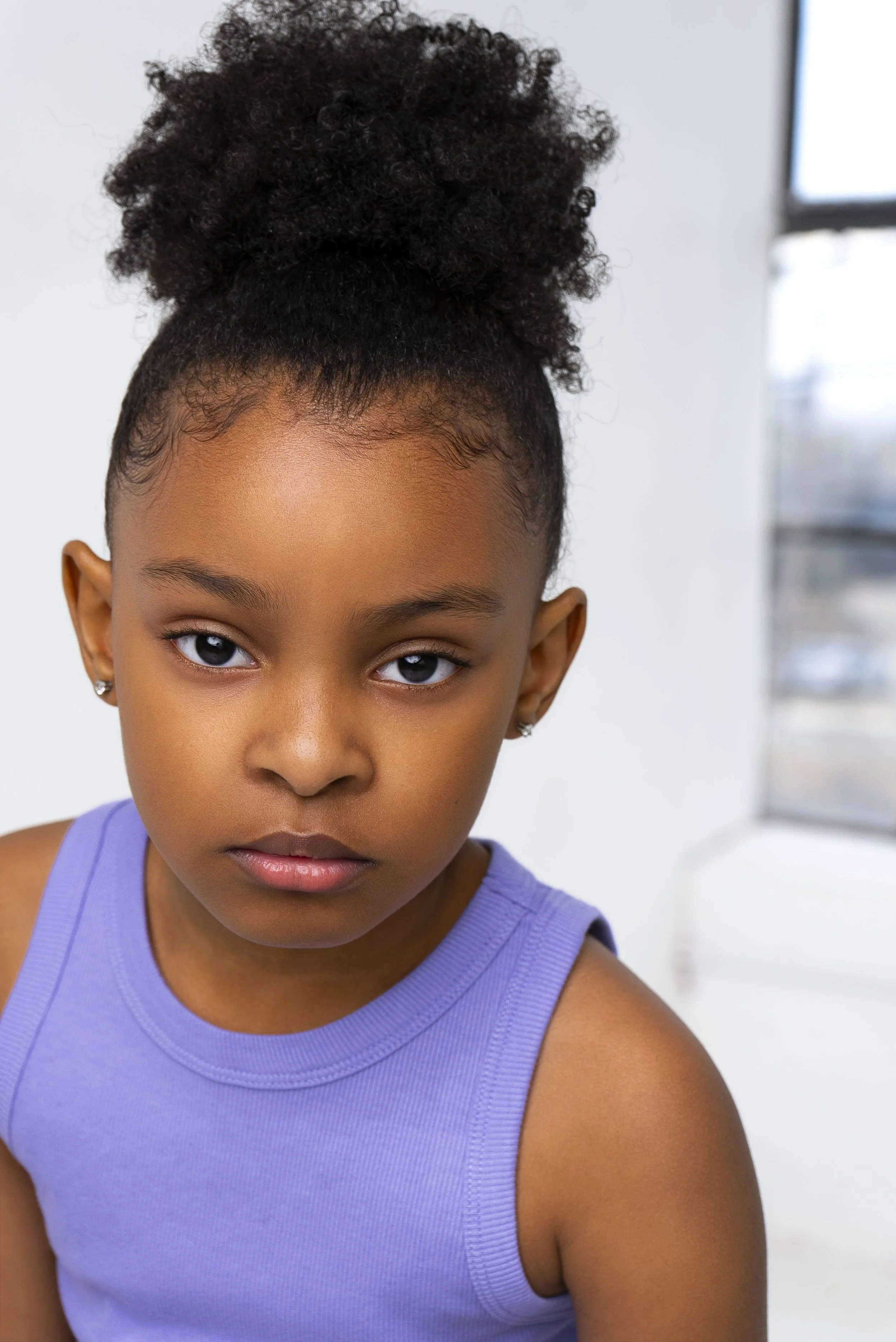 A young girl with dark skin, wearing a purple sleeveless top, has neatly styled curly hair in a high puff, and is looking directly at the camera with a serious expression.
