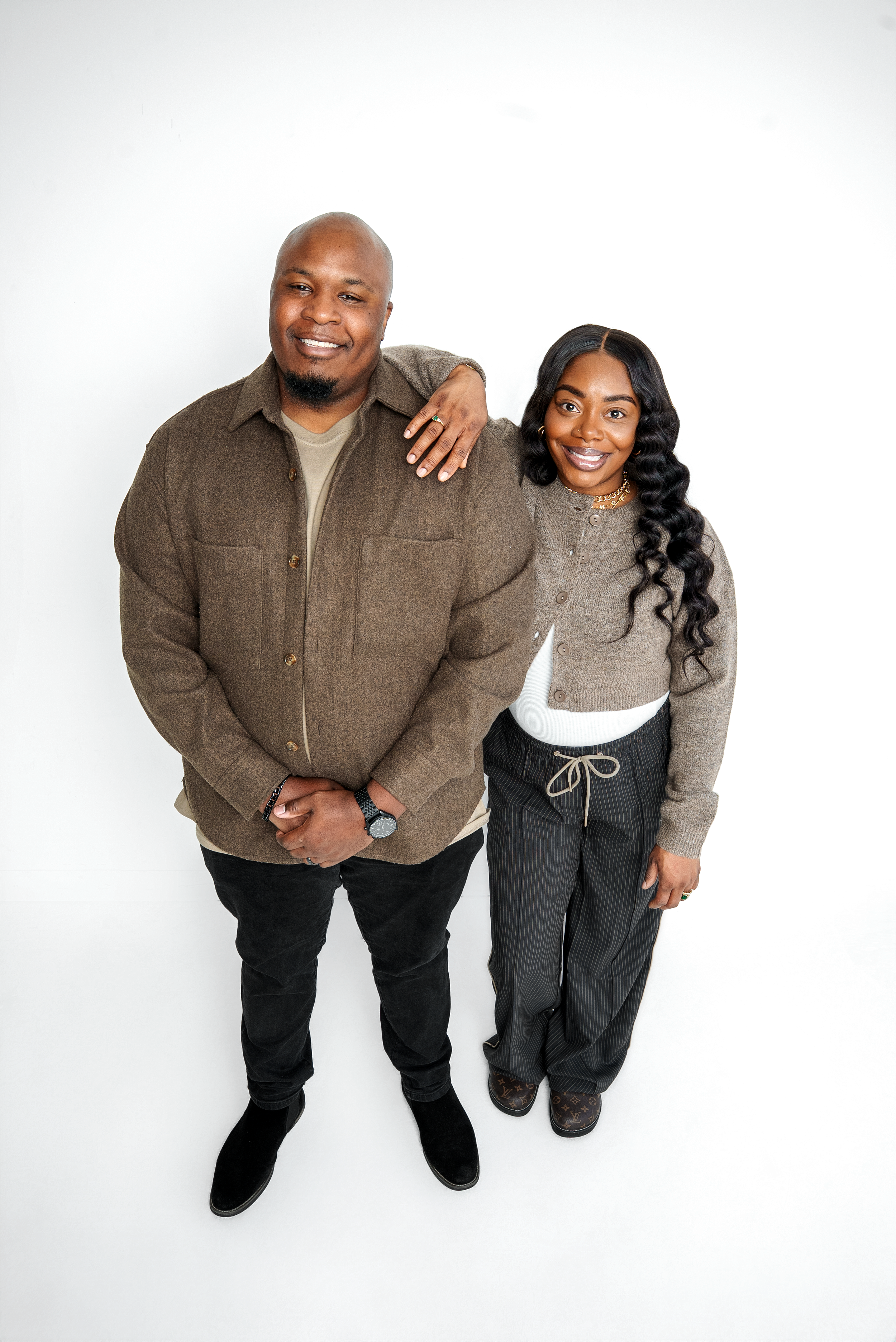 A smiling man and woman standing together against a white background. The man is wearing a brown jacket, black pants, and black shoes, while the woman is dressed in a beige cardigan, white top, pinstriped pants, and designer shoes. The woman has her 