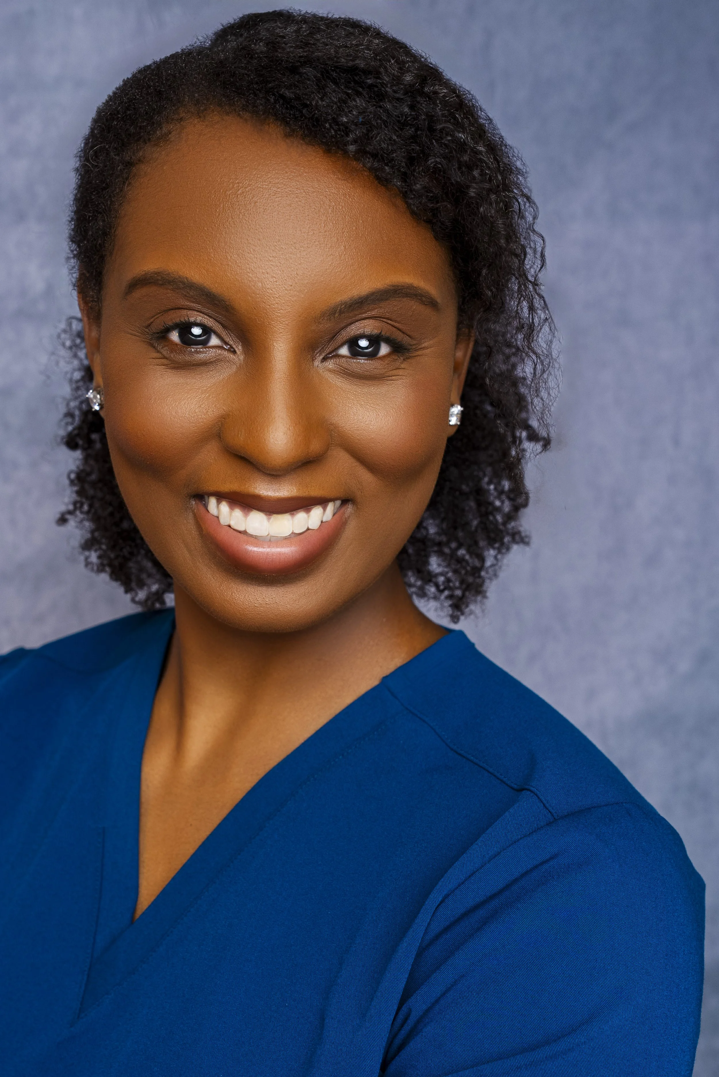 Smiling African American woman with curly hair, wearing blue scrubs, earrings, and makeup, posing against a gray background.