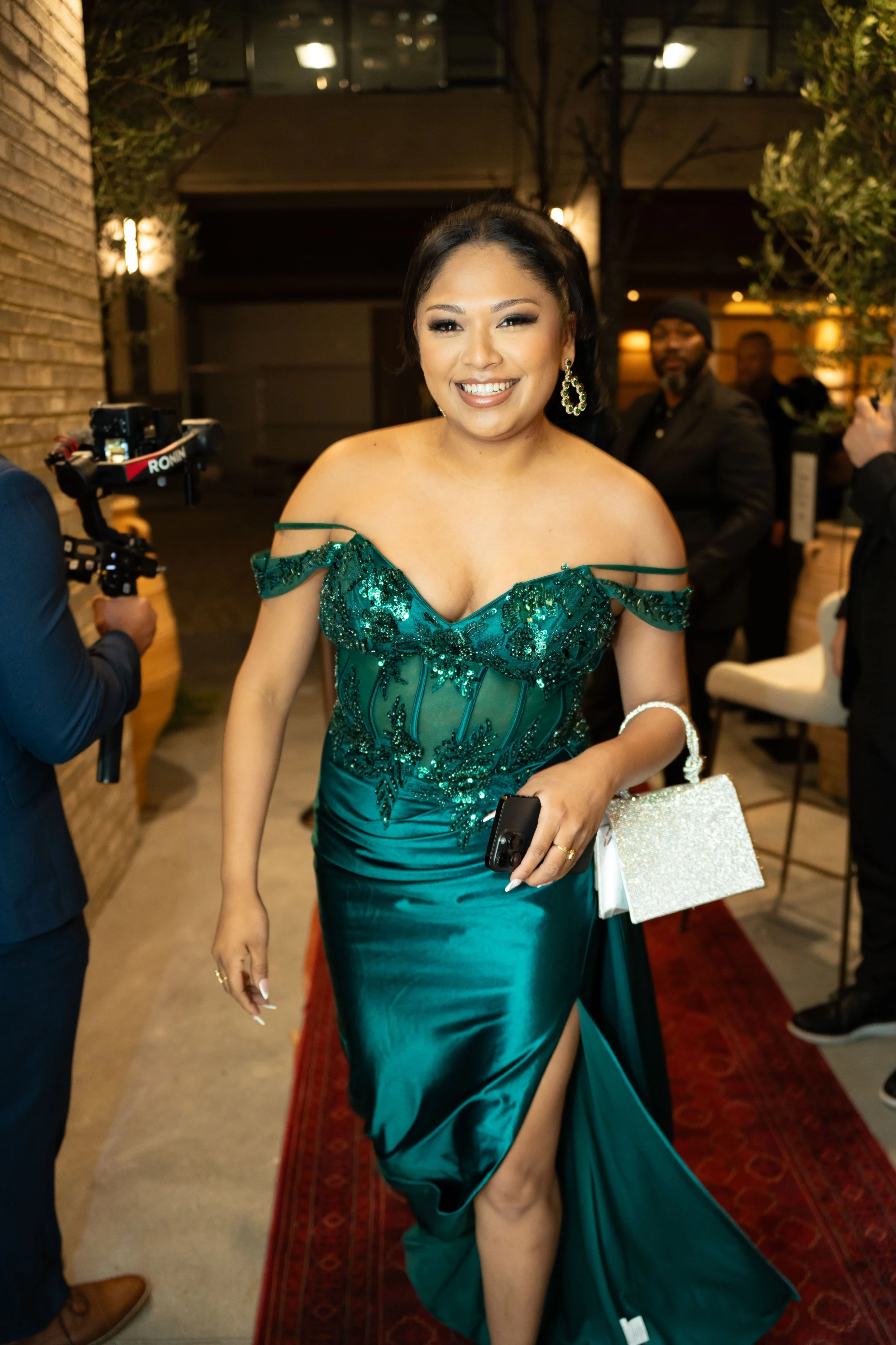 Woman in a teal, sequined evening gown walking on a red carpet at an event, holding a white handbag and a phone.