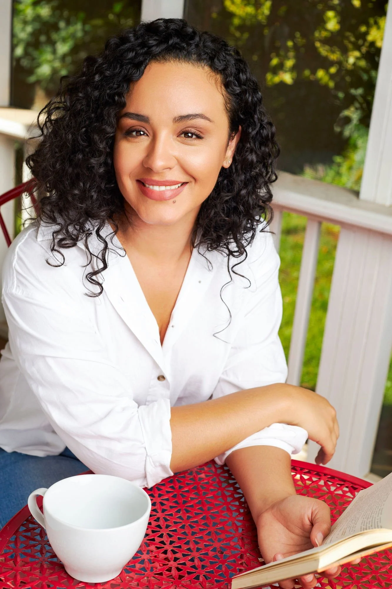 A woman with curly black hair and a white shirt sitting at a red table outdoors, smiling, with a white mug nearby and reading a book.