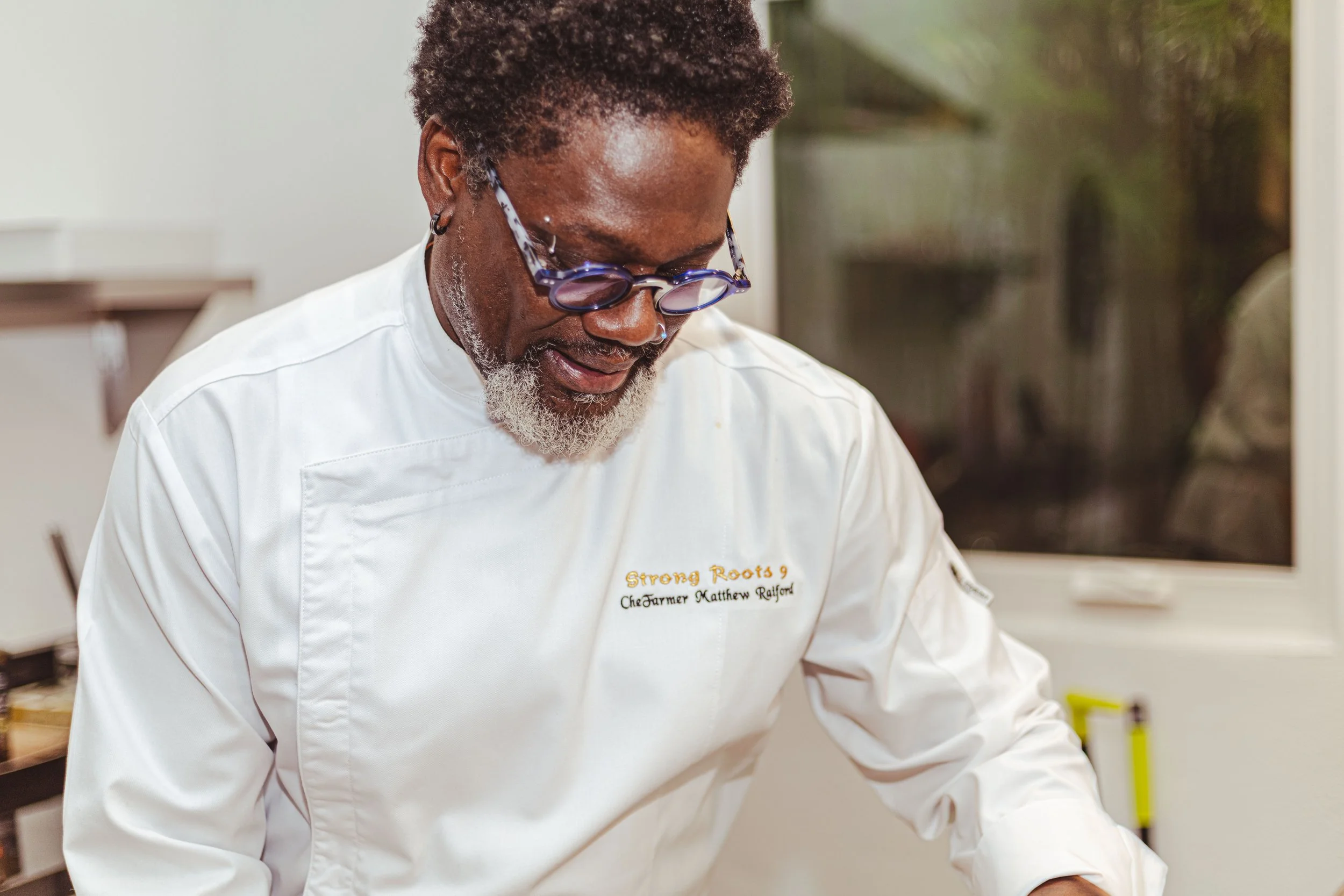 A chef with glasses and a beard, wearing a white chef's coat, working in a kitchen.