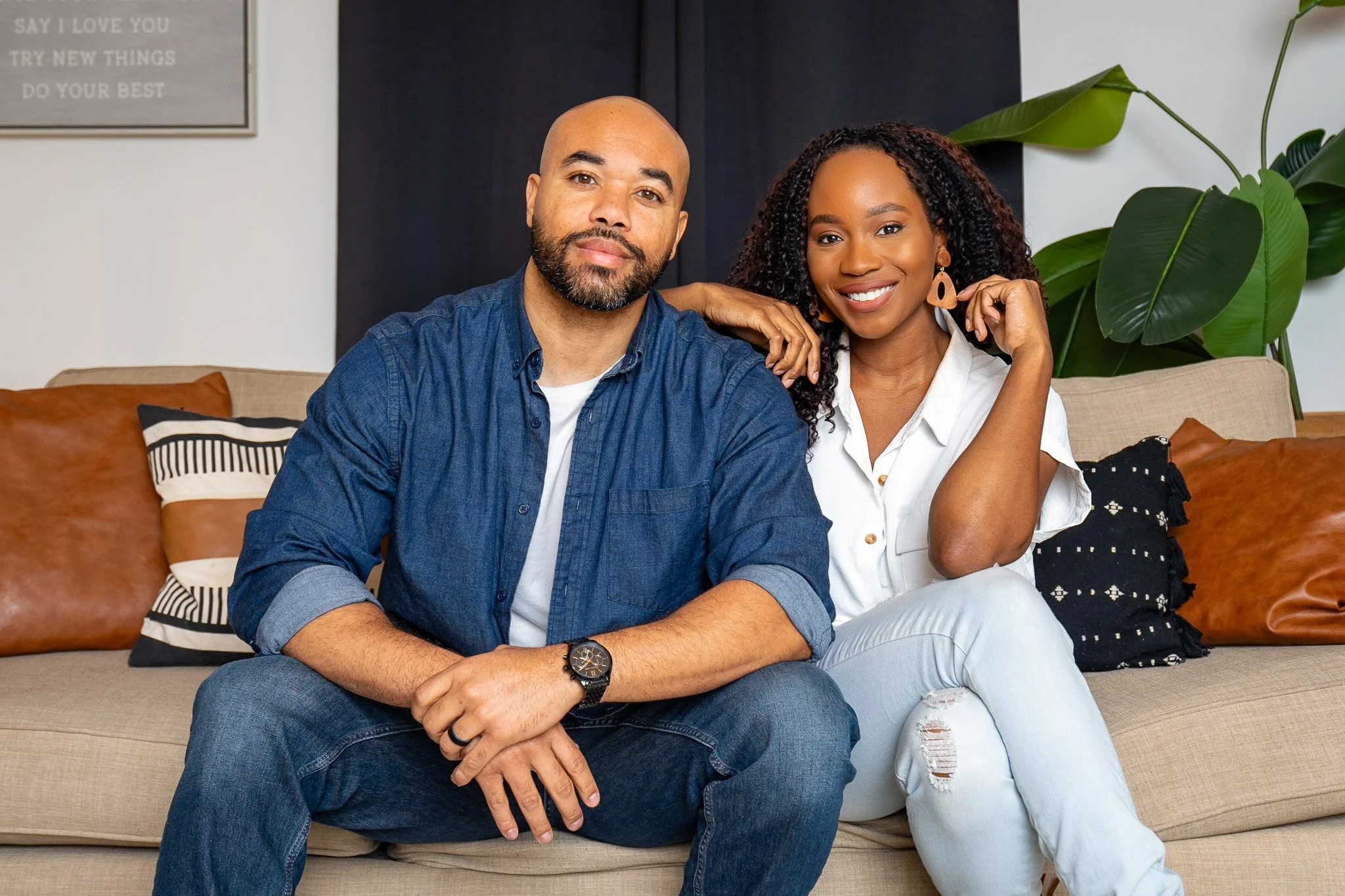 A man and a woman sitting on a beige sofa in a living room, smiling at the camera. The man has a beard, is wearing a blue denim shirt and jeans, and has a watch on his left wrist. The woman has curly hair, is wearing a white blouse, ripped jeans, and large earrings. There are decorative pillows on the sofa, and a large plant with broad leaves is in the background. There is a framed quote on the wall in the background.