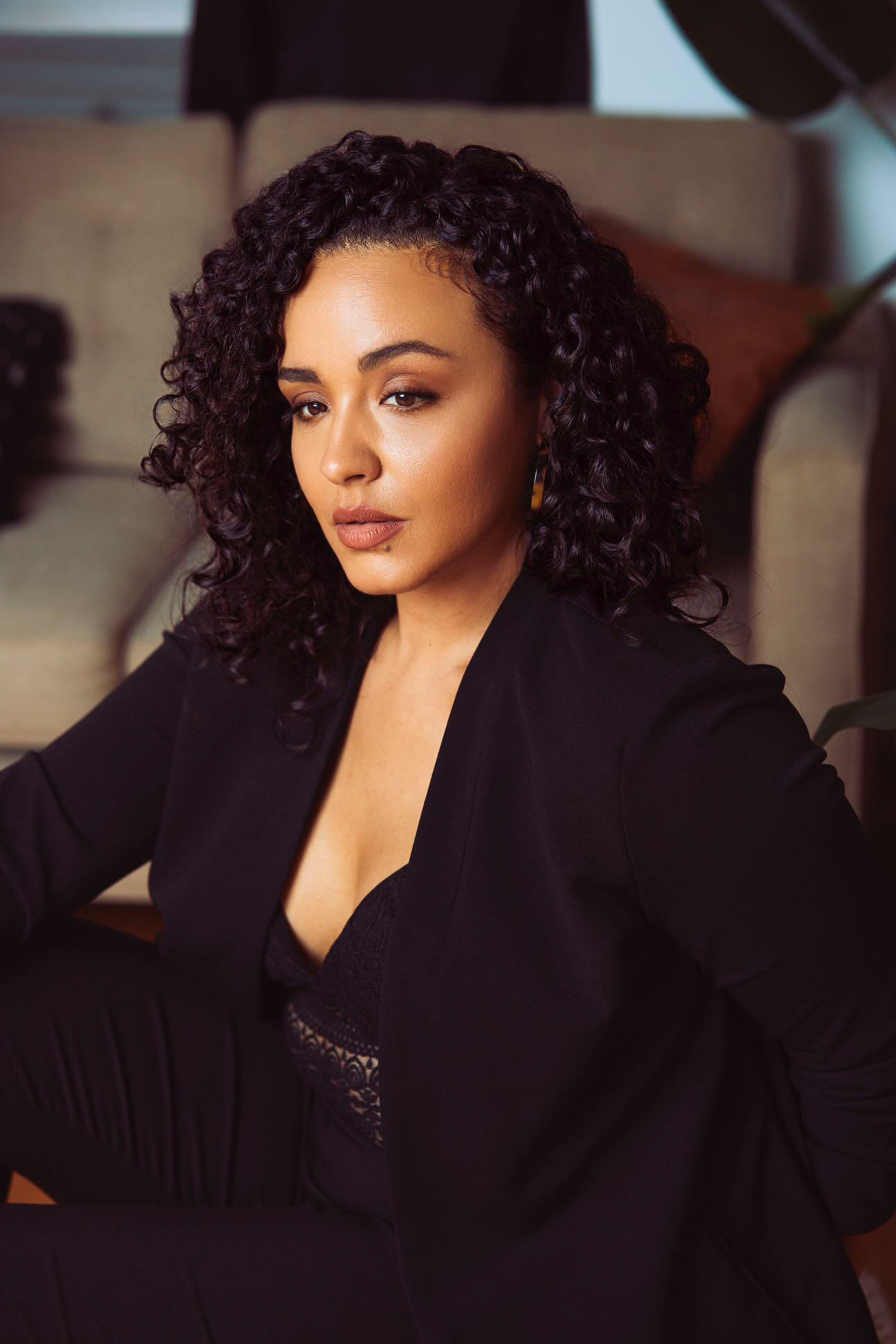 A woman with curly dark hair, wearing a black blazer and black top, seated indoors in what appears to be a living room setting.