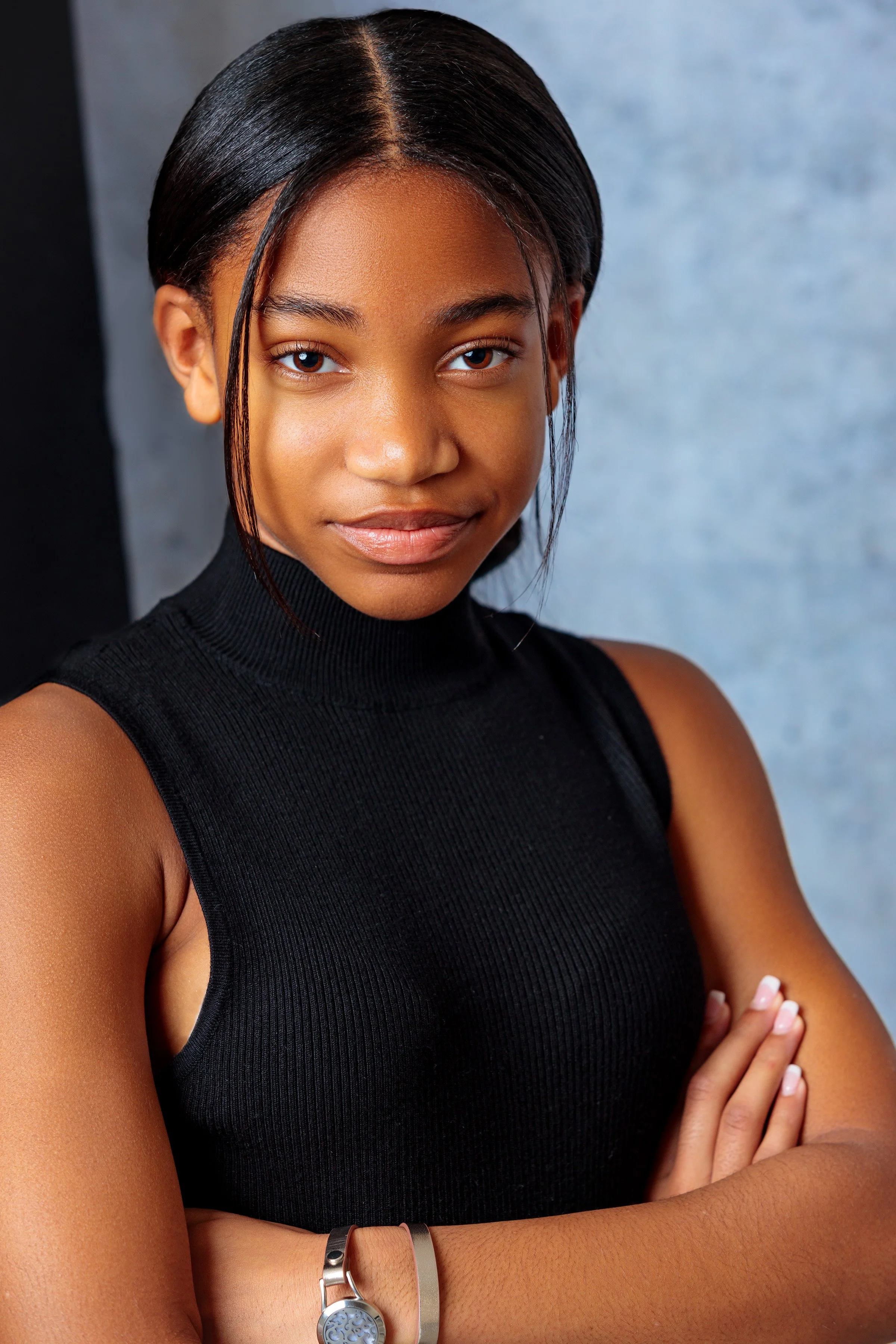 Close-up portrait of a young woman with dark hair and brown skin, wearing a sleeveless black turtleneck, standing against a blurred background, with her arms crossed and looking at the camera.