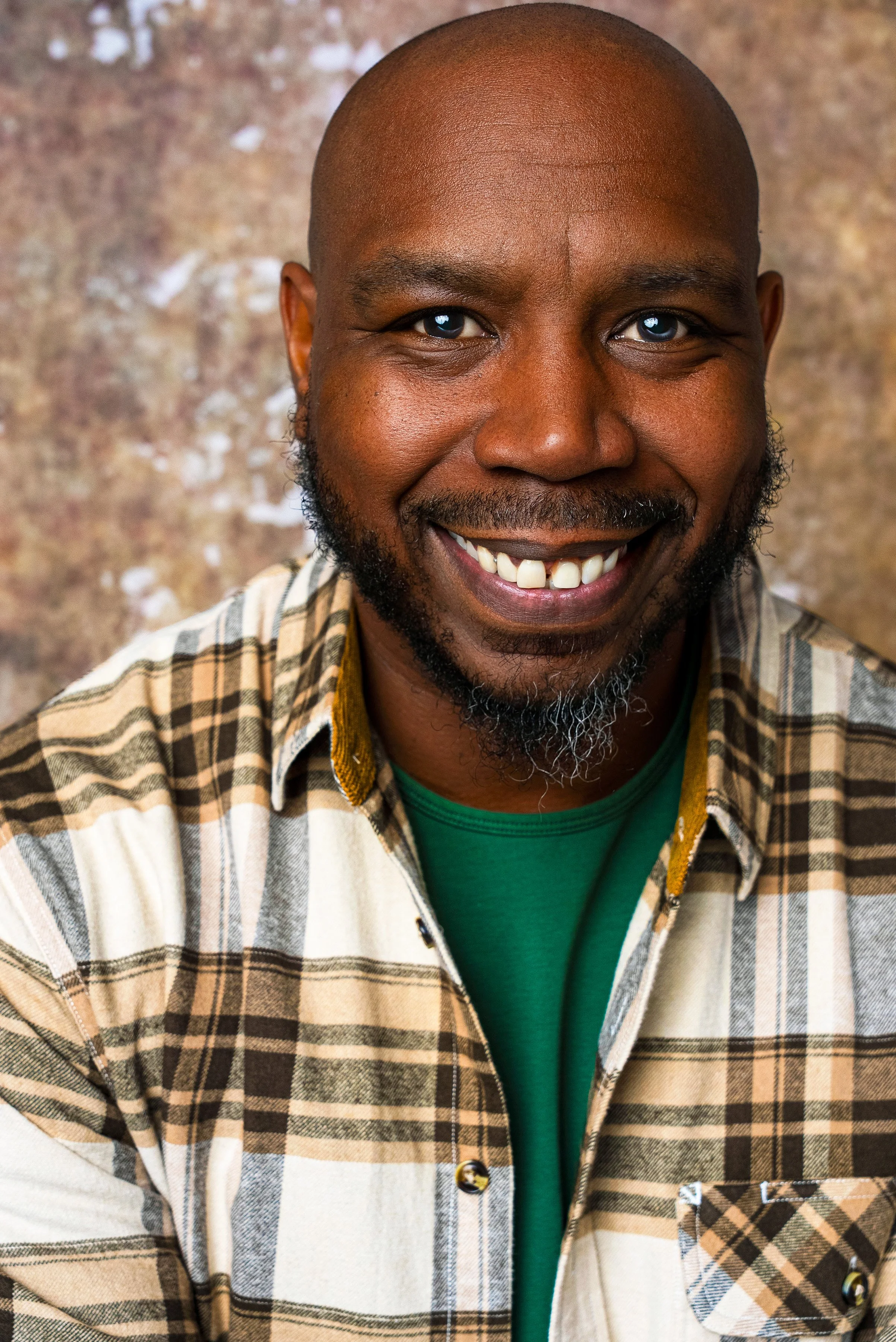 A smiling man with a beard and mustache wearing a plaid shirt over a green t-shirt, with a textured brown background.