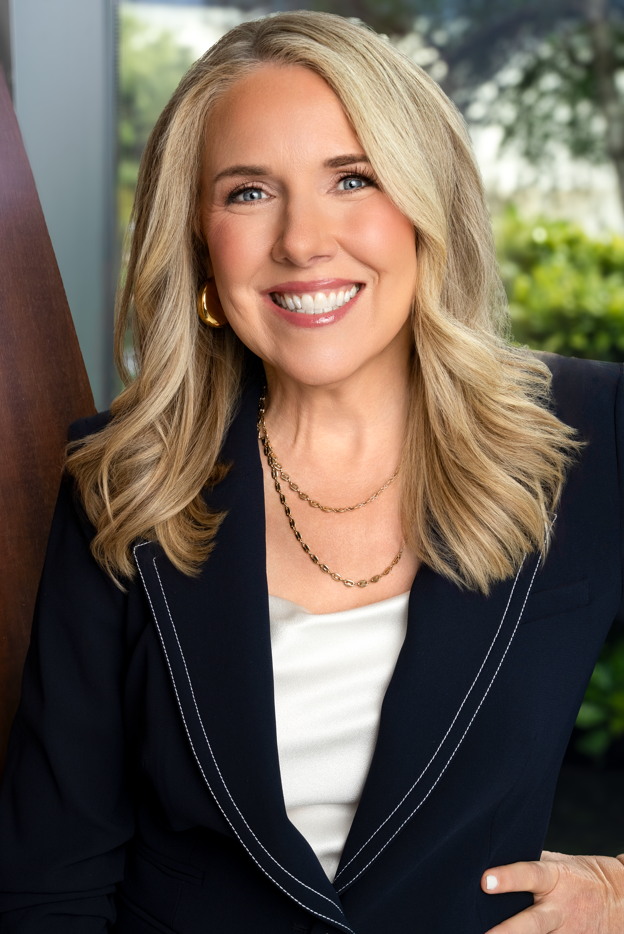 A smiling woman with blonde hair, wearing a dark blazer over a white top, gold jewelry, and seated outdoors with greenery in the background.