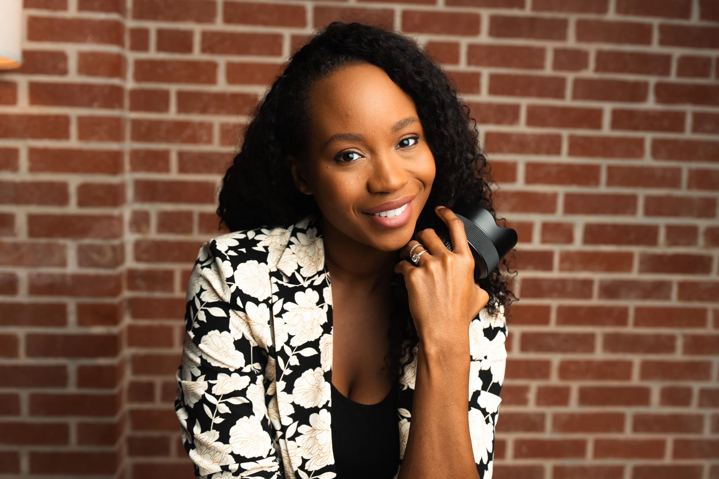 A smiling woman with curly hair holding a camera, wearing a black and white floral jacket, standing in front of a brick wall.