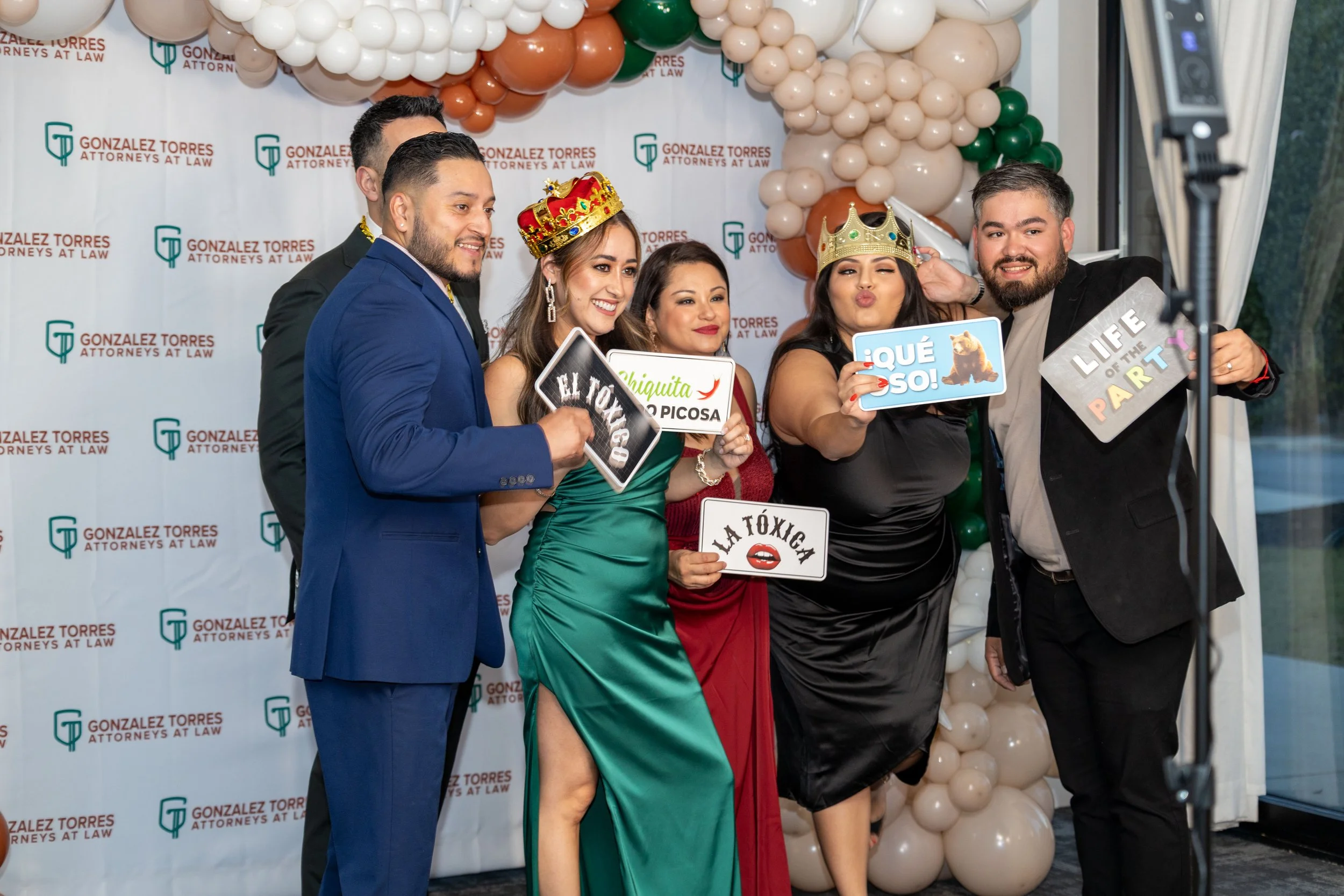 Group of six people celebrating at a formal event with balloons and a backdrop that reads "Gonzalez Torres Attorneys at Law." Some individuals wear crowns and hold photo booth signs with congratulatory messages.