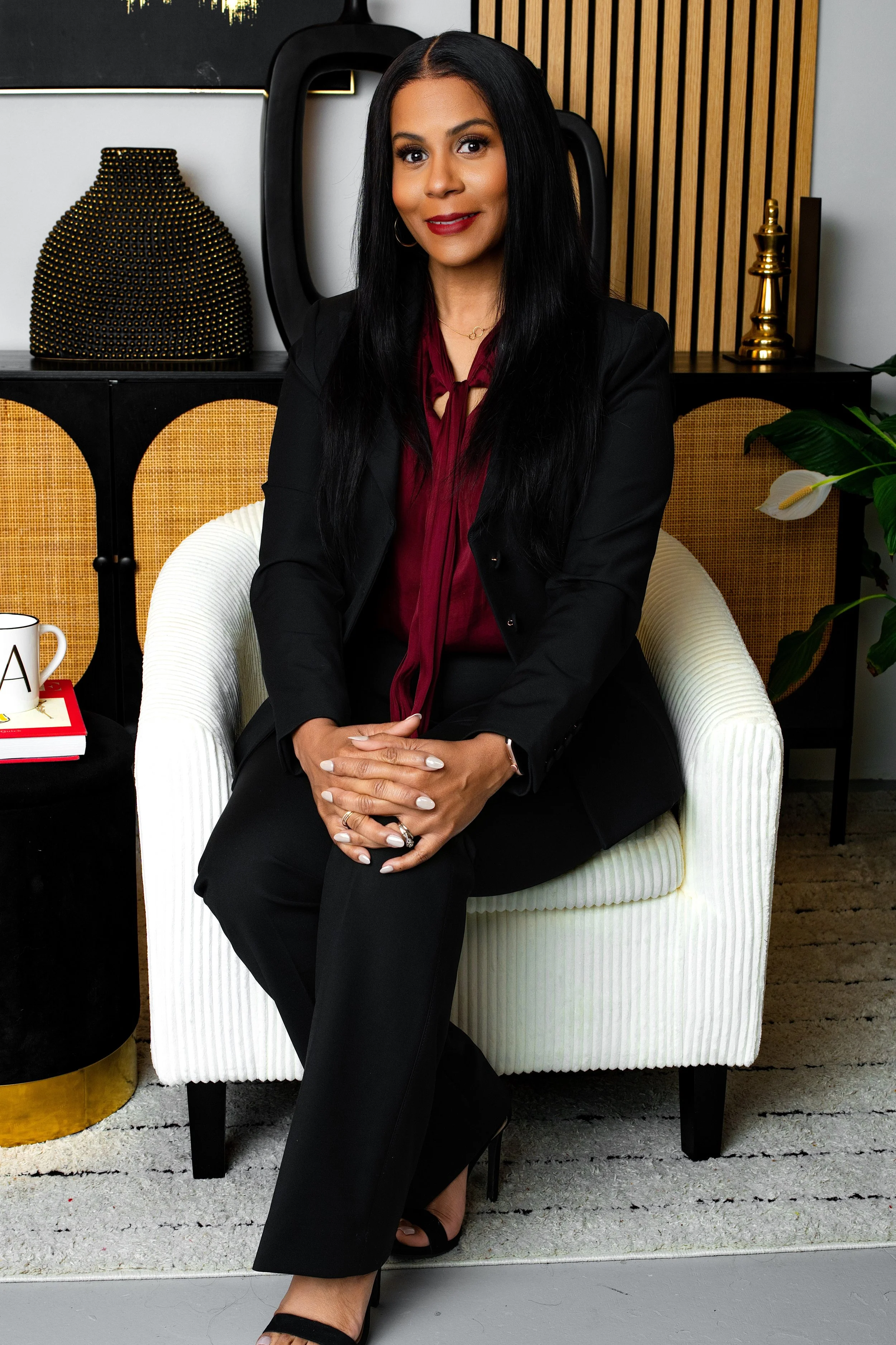 A woman sitting on a white armchair with a black sleeve blazer and burgundy blouse, with a modern living room background including a black table, a black and gold decorative lamp, a mug, and a stack of books.