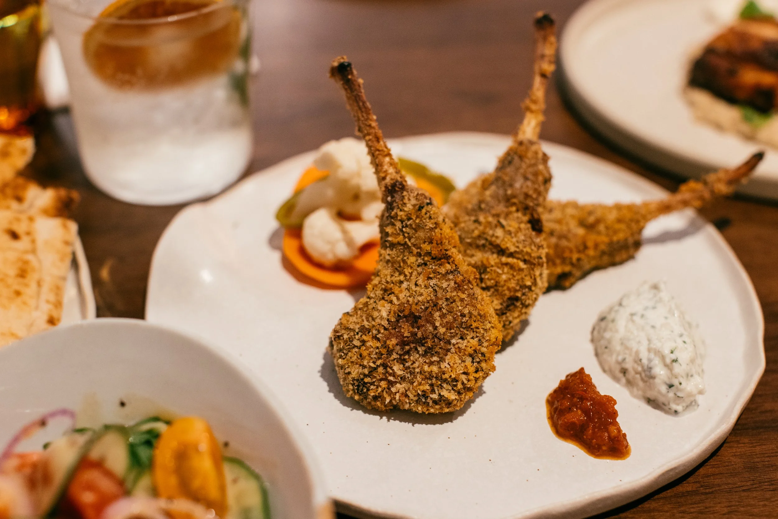 Breaded lamb chops served with tzatziki and harissa on a white plate, with chunky homemade pickles and a gin & tonic in the background.