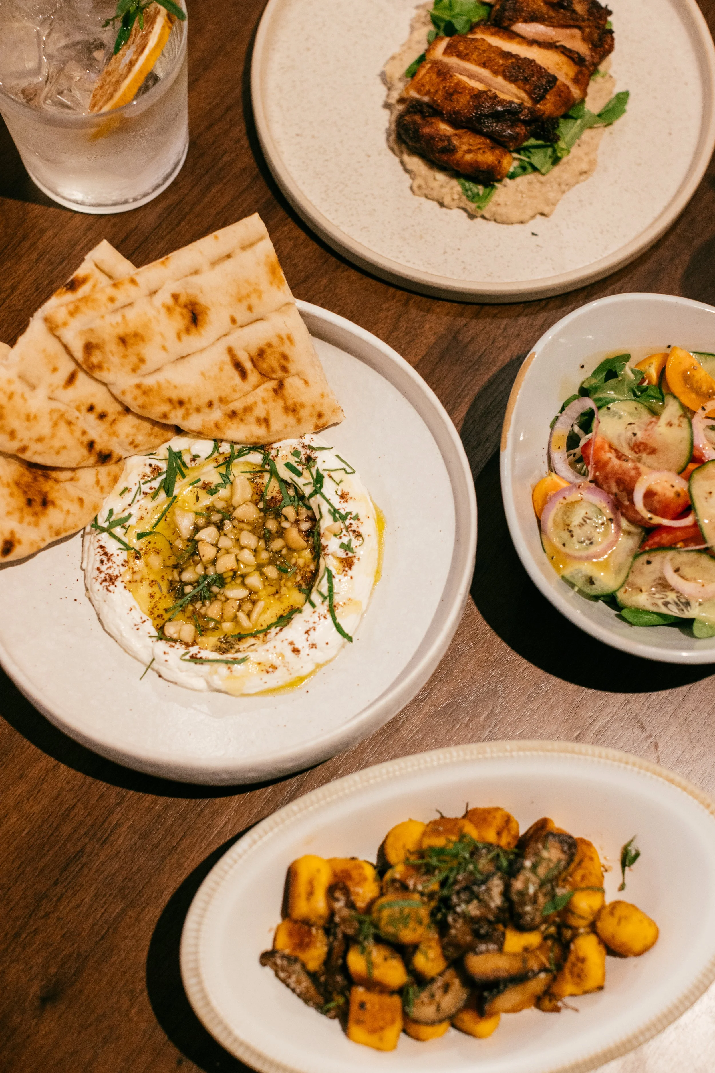 A variety of Middle Eastern dishes. Labneh topped with nuts & honey, grilled meat, a fresh salad with cucumbers, tomatoes, and onions, and pan-fried gnocci topped with herbs. There is also a glass of vermouth-soda with a dried grapefruit and ice.