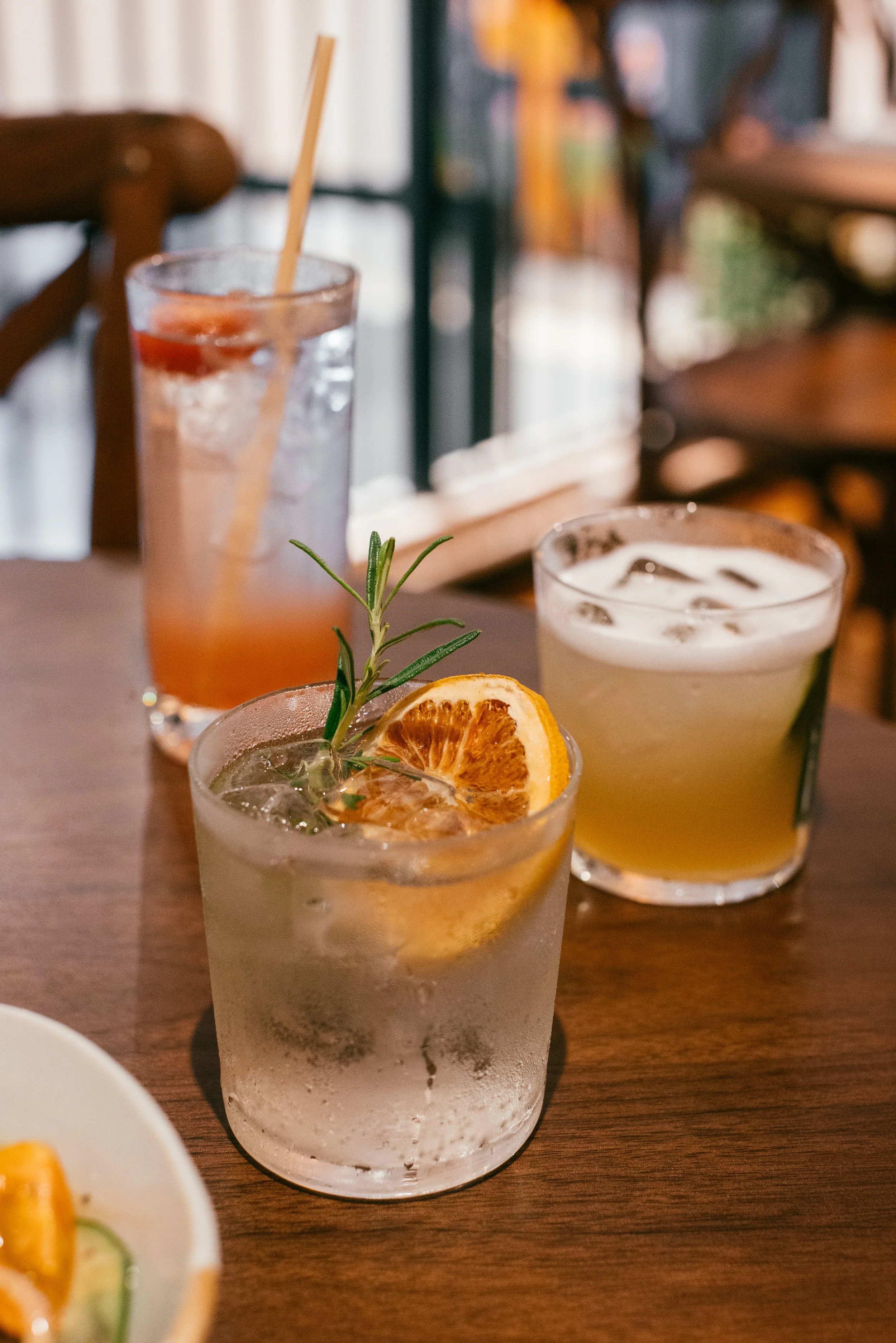 A selection of cocktails including cucumber sour, watermelon and cherry tomato gin fizz and a vermouth soda with dried grapefruit and rosemary, placed on a wooden table with other drinks in the background at a restaurant or cafe.
