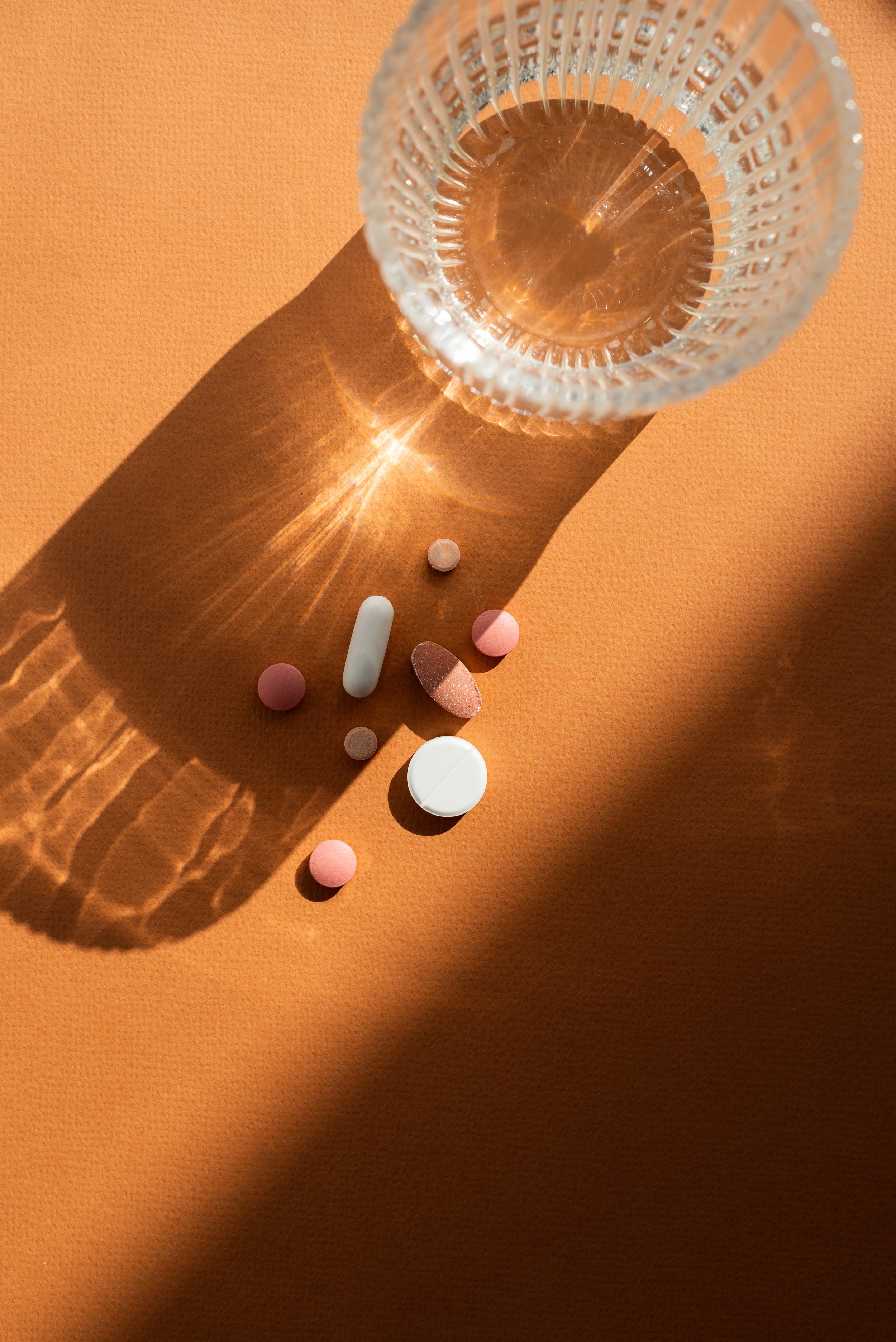 A glass of water placed on an orange surface with pills and tablets scattered underneath, casting shadows and reflections.