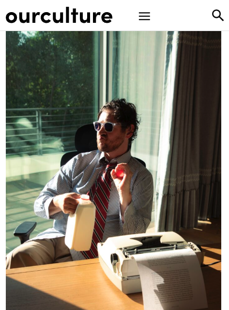 A man with sunglasses, a striped shirt, and a tie, sitting at a desk with a typewriter, holding a milk carton in one hand and an apple in the other, in front of a large window with sunlight and curtains.